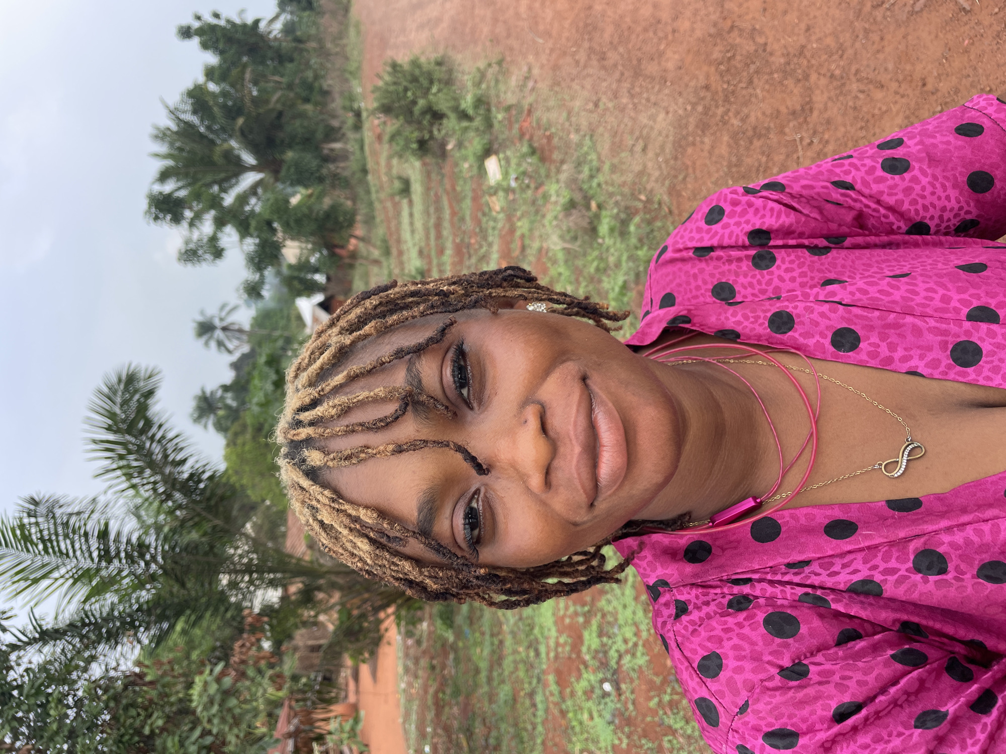 A medium close-up shot shows a woman smiling at the camera in an outdoor environment with tropical vegetation. She is wearing a bright pink shirt with black polka dots and two necklaces, one of which has an infinity symbol pendant. Her hair is styled in dreadlocks that are dark brown near the scalp and blonde at the tips. The background shows a dusty, reddish-brown ground with patches of green vegetation and trees, with the overcast sky visible at the top. This image likely was taken in Ogbe, Nigeria.