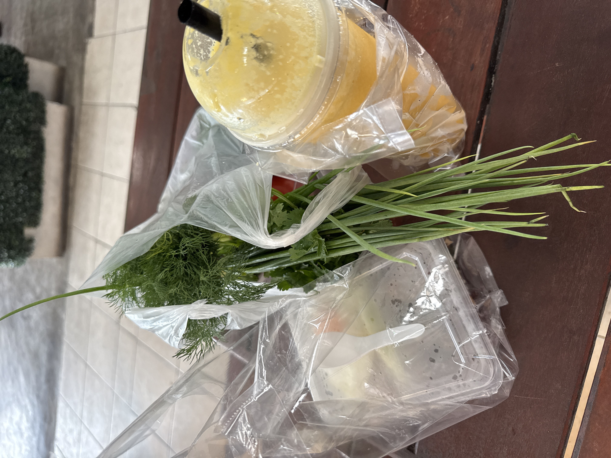 This overhead, high-angle shot captures a collection of food and drink items placed on a dark wooden table. In the upper portion of the frame, a clear plastic cup filled with a thick, yellow beverage is visible, topped with a black straw. The cup is partially enclosed in a clear plastic bag. Below this, a larger clear plastic bag contains fresh green herbs, including dill and scallions. To the right of the herbs, a clear plastic container with a white plastic utensil inside holds a portion of food, possibly a salad or side dish. The overall scene suggests a casual meal or takeout order. The lighting appears to be natural, indicating daytime. The backdrop includes a blurred tiled surface on the left and the dark wood grain of the table. No visible text is present in the image. The setting appears to be an outdoor or semi-outdoor space, possibly a patio or balcony, given the faint background details that could be a railing or steps. The overall mood is relaxed and informal.