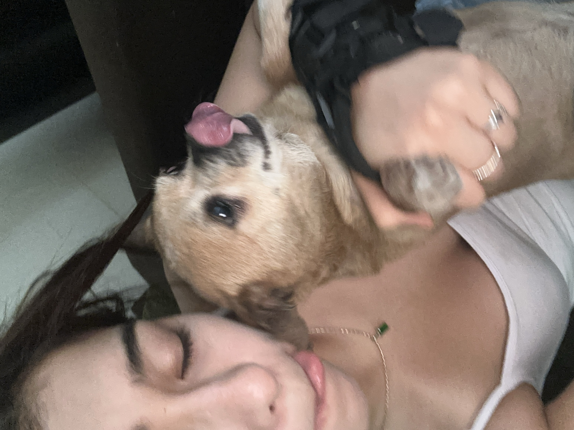 The image captures an intimate, close-up moment indoors between a person and a small, light brown dog. The person, appearing to be a woman, is seen from the nose up, with her eyes closed and a relaxed expression, as the dog licks her face, specifically around her mouth and chin. Her dark hair falls around her head. She wears a white tank top and a delicate gold-colored necklace with a small green pendant. A hand, likely hers, is visible holding one of the dog's paws or legs, adorned with two rings: one silvery with a dark stone, and another a ribbed band. The dog, positioned above the person, has its pink tongue extended during the lick, and a dark accessory, possibly a harness or vest, is visible on its back/shoulder area. The background is dark and indistinct, suggesting a dimly lit room or a very close crop. The overall mood conveyed is one of affection, playfulness, and comfort between the pet and its owner. While the location context is given as Puebla, Mexico, there are no specific visual cues within the image to confirm this. There is no visible text.