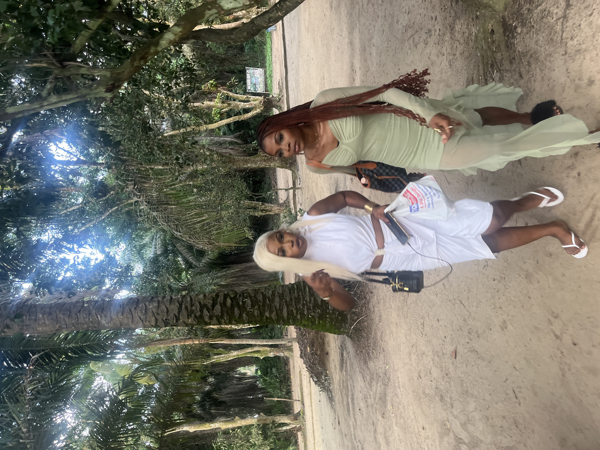 Two women walk on a sandy path in a tropical environment, possibly a resort in Lekki, Nigeria. The woman on the left has blonde hair and wears a white ruched dress with a cutout at the waist. She carries a black purse with a gold clasp and a white plastic bag. The woman on the right has long, braided auburn hair and wears a light green sheer dress with ruching and a ruffled hem. She carries a checkered patterned bag. Both women are wearing open-toed footwear. The background features dense foliage, including palm trees, with a sign visible in the distance. The overall lighting suggests it is daytime, with sunlight filtering through the trees.