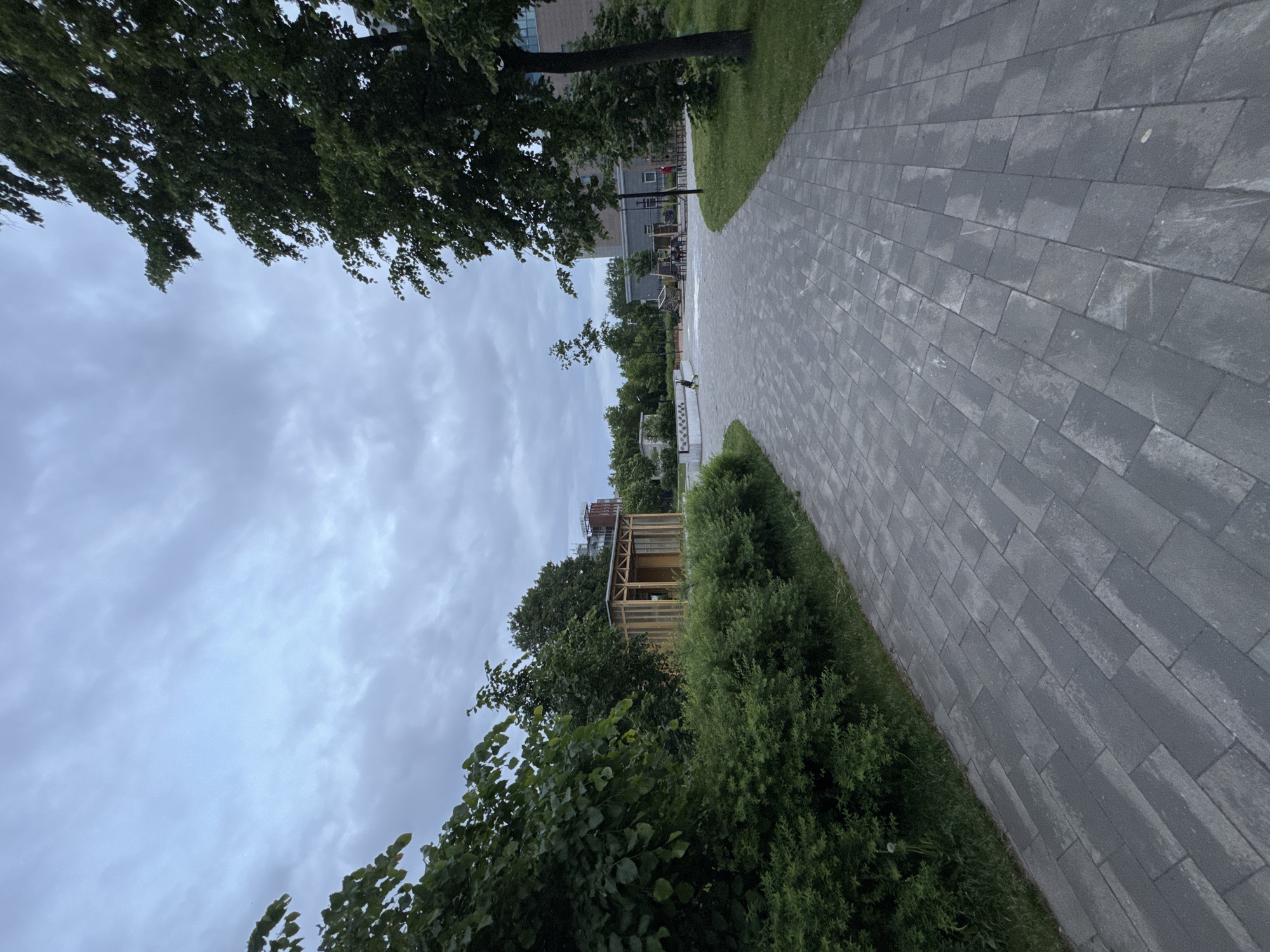 The image displays an outdoor, landscaped urban environment, likely a park or courtyard, under a heavily overcast sky. A wide pedestrian pathway, constructed from grey rectangular concrete pavers, extends from the foreground into the mid-ground, subtly curving towards the right. The path appears somewhat damp or discolored in patches.
Dense green foliage, comprising both low-lying bushes and mature trees, lines both sides of the pathway. On the left, nestled behind a thick hedge, is a contemporary wooden structure, possibly a kiosk or shelter, characterized by a light-colored timber frame, slatted roof elements, and large vertical panels that appear to be glass. Further in the background on the left, above the treeline, a multi-story building with a reddish-brown upper section is visible, bearing the Cyrillic text "СВОГРА" (SVOGRA) on its facade. This sign identifies the location as Izhevsk, Russia, where "SVOGRA" is a known construction company.
On the right side of the path, a manicured lawn separates the paving from a line of tall, leafy trees. Beyond these trees and further into the mid-ground, a children's playground with various equipment is visible, situated in front of a larger, multi-story building with a light-colored brick and grey facade. Several indistinct human figures, consistent with adults and children, are present near the playground. The entire sky is covered by a uniform layer of grey, textured clouds, indicating an overcast day with soft, diffused lighting.