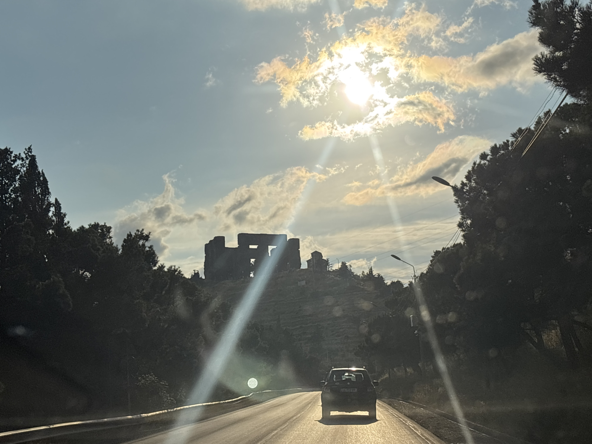 The image captures a view from inside a vehicle driving on a road in Tbilisi, Georgia. The road curves to the left, with a dark guardrail visible along the right edge. On the right side of the road, dense trees line the background, with a streetlamp visible amongst them. In the distance, perched atop a hill, are the ruins of a stone structure, appearing somewhat blocky and dilapidated, with some smaller buildings visible near it. The sun is bright in the sky, casting strong lens flares and light rays across the image, illuminating scattered clouds that are tinged with yellow and orange. The sky is a pale blue. A dark-colored SUV is driving ahead on the same road, its license plate partially visible. The overall scene suggests a sunny day, with the vehicle traveling through a scenic area with historical ruins.
