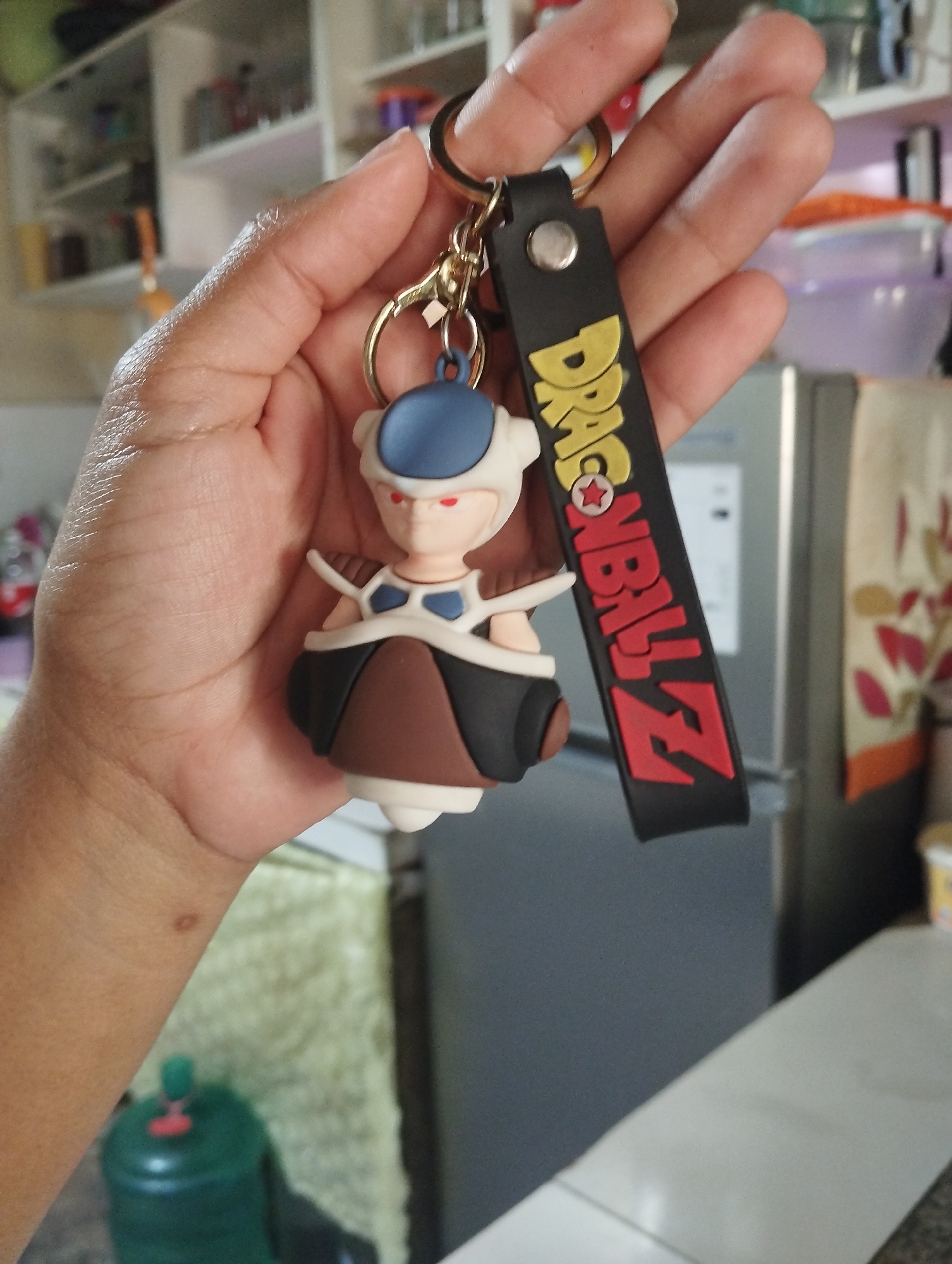 A hand is holding a Dragon Ball Z keychain. The keychain features a small figurine of a character with a blue helmet, red eyes, and a brown and black outfit, attached to a black strap with the Dragon Ball Z logo in yellow and red. The background is blurred, showing shelves filled with various items and a gray refrigerator. The scene appears to be indoors, possibly in a kitchen or storage area, and is located in El Valle del Espíritu Santo, Venezuela.