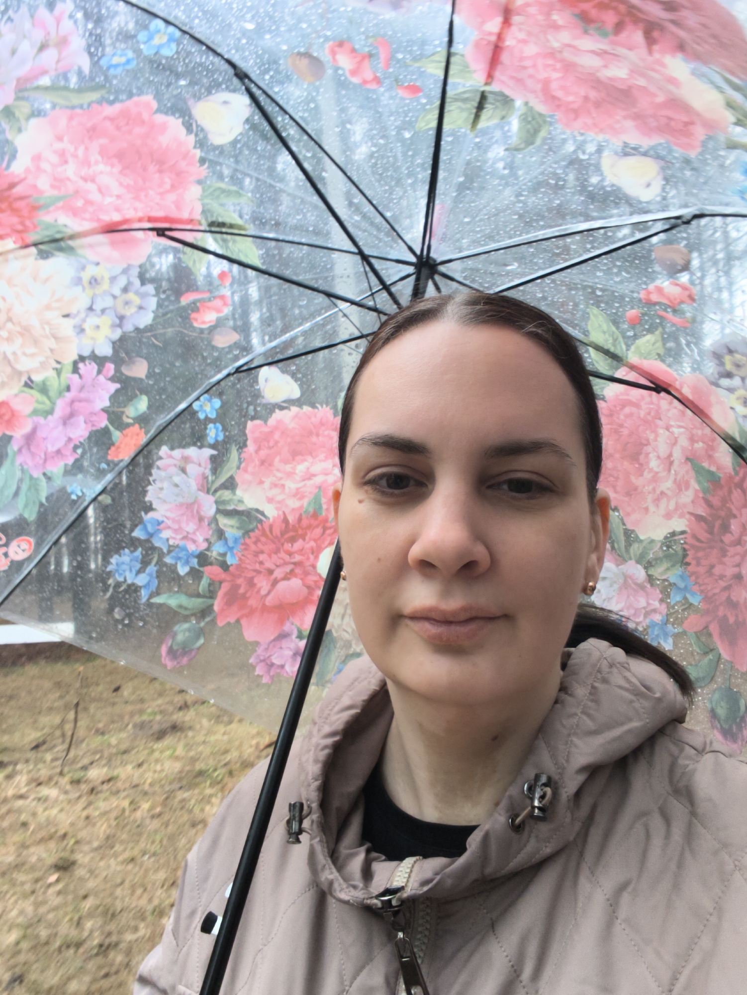 A woman is captured in a headshot-style selfie, holding a clear, transparent umbrella with a vibrant floral pattern. She is shown from the chest up, looking directly at the camera with a neutral expression. Her dark hair is pulled back, and she has light skin and brown eyes. She is wearing a light beige or tan quilted jacket over a dark-colored top.
The prominent umbrella, held above her head, features large illustrations of flowers, predominantly in shades of pink and red, with accents of blue, purple, and green foliage. The surface of the umbrella is covered in numerous small raindrops, indicating rainy or recently rainy weather in Chaikovsky, Russia. Through the clear, rain-dotted umbrella, an indistinct outdoor background is visible, consisting of brown ground cover, possibly wet earth or dormant grass, and subtle hints of distant trees or structures. No specific landmarks are discernible. There is no visible text in the image.