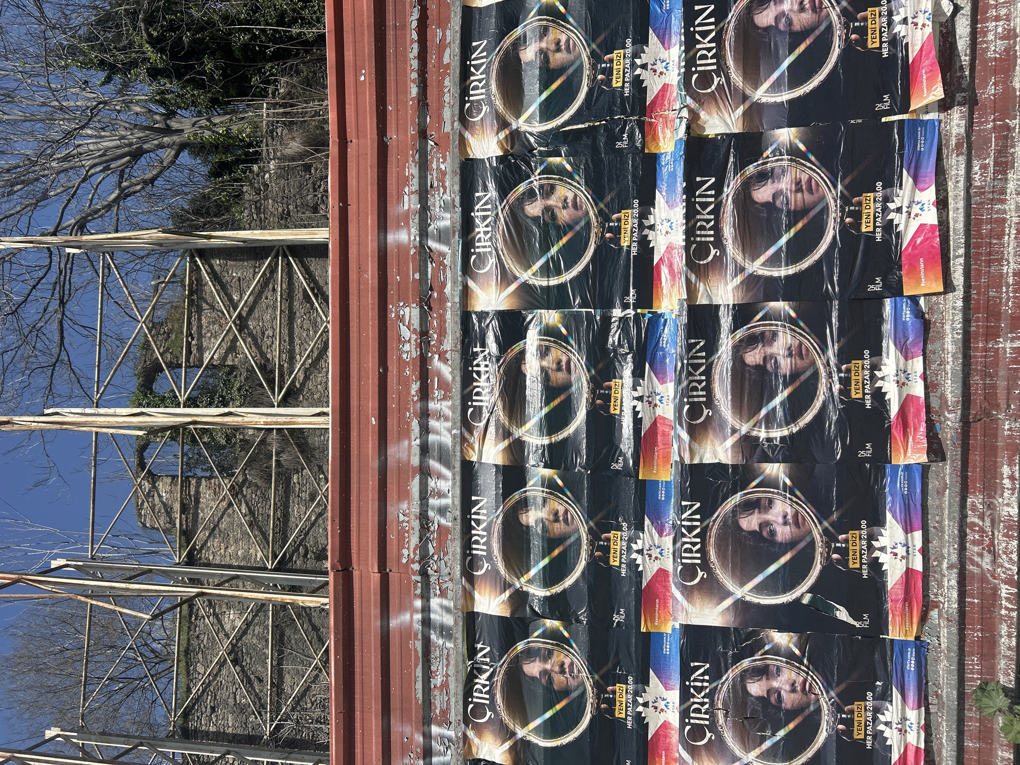 The image depicts a series of posters for a Turkish television show called "Çirkin" (meaning "Ugly") affixed to a weathered, corrugated metal wall. The posters feature a close-up of a person's face within a circular frame, overlaid with a prism effect. Each poster also includes the text "YENI DIZI HER PAZAR 20.00" (New series every Monday 8:00 PM), along with other text and branding including "Star" and "#seninstarın" (your star).
To the left of the posters, there is a structure made of metal beams and supports, and the sky is visible through gaps in the construction. There's also a tree with bare branches visible above the structure. The building looks weathered and is made of stone.
The image orientation is tilted, possibly taken while holding the camera sideways. Given the "startv.com.tr" branding on the posters, and the reference to Beyoğlu in the prompt, it's plausible the posters are located in this city district in Istanbul, Turkey.