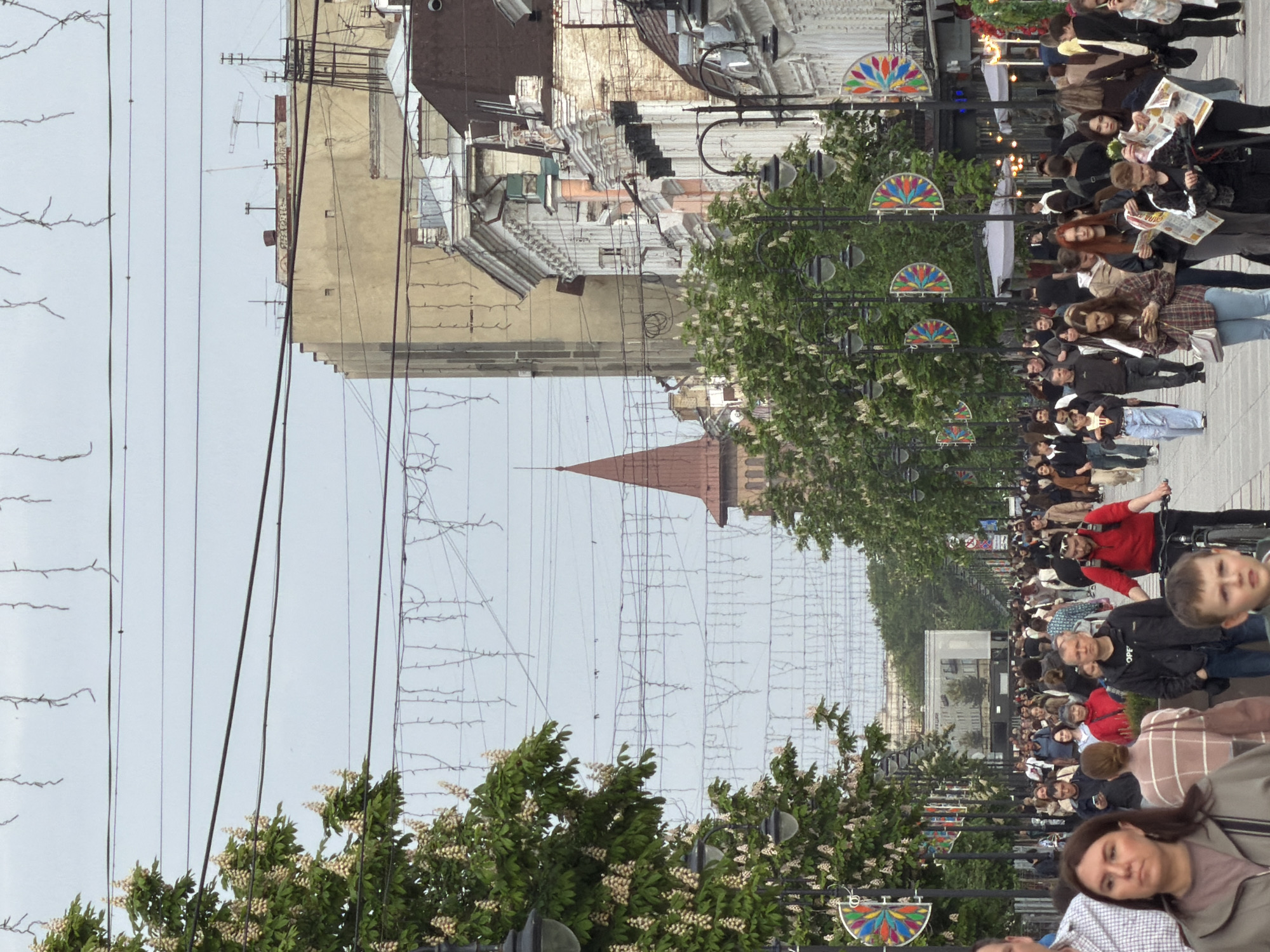 The image depicts a crowded street in Saratov, Russia, viewed from an elevated perspective along the street's axis. People are walking along a paved sidewalk lined with trees, streetlights decorated with colorful, semi-circular ornaments, and overhead wires. Buildings of varying heights and architectural styles are visible on both sides of the street. A pointed, red tower rises above the buildings in the middle distance. Some people are looking at newspapers. The sky is overcast.