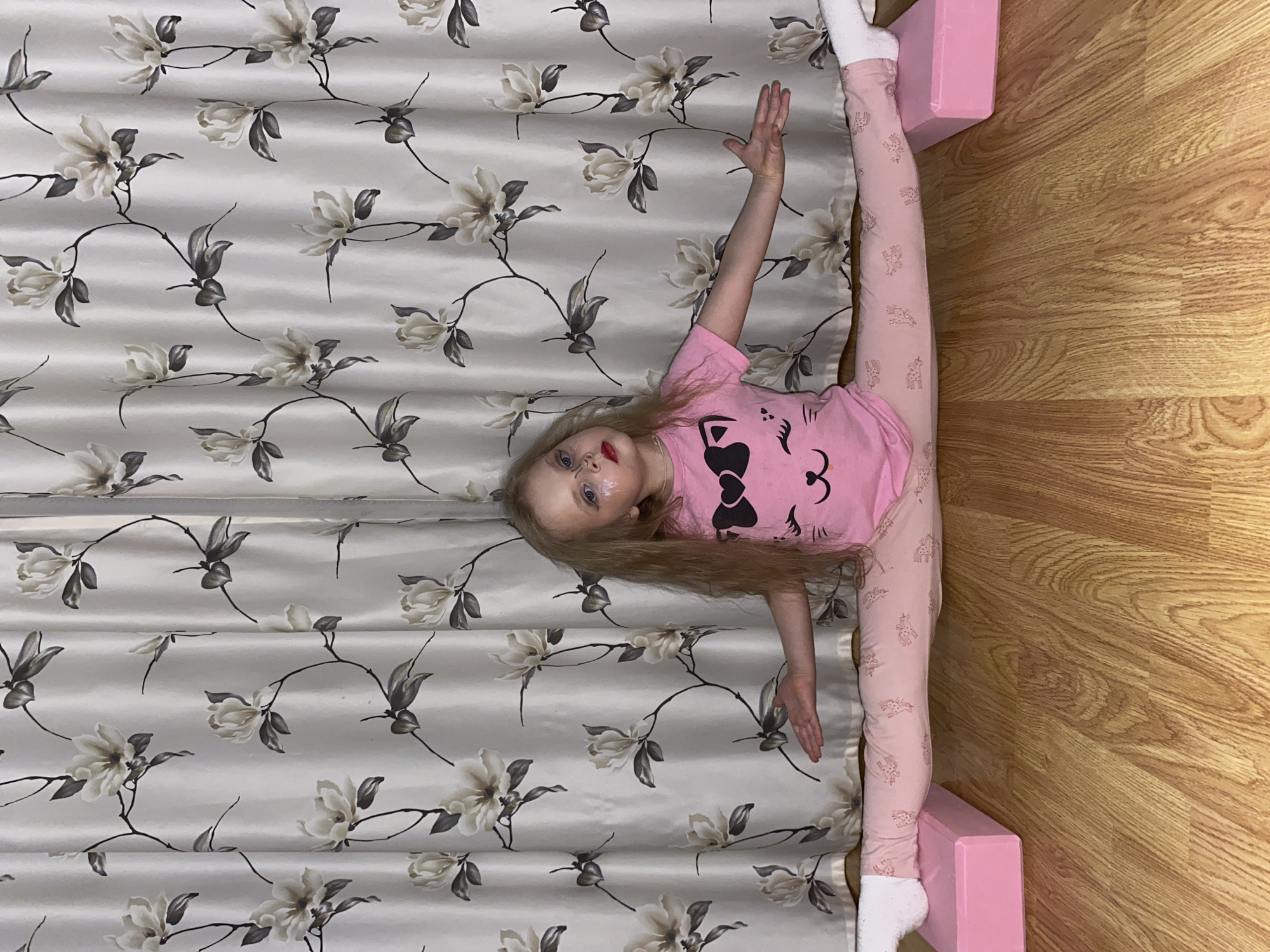 A young child is captured in a vertical, overhead shot, performing a full split across two distinct floor surfaces. The child, with long blonde hair, is wearing a pink short-sleeved t-shirt featuring a black bow design and pink leggings patterned with what appears to be small unicorns or similar figures. White socks cover her feet. She also has some visible makeup, including red lipstick.
Her body is positioned horizontally across the frame. Her left leg extends onto a light-colored surface patterned with delicate white/beige flowers, dark leaves, and slender branches, resembling a floral curtain or a soft mat. Her right leg stretches onto a light brown wooden floor, showcasing prominent wood grain, with her right foot resting on a pink rectangular block. Her left arm is bent upwards above her head, while her right arm rests by her side, hand open. The child looks towards the camera with a focused expression.
The setting appears to be indoors, likely a home in Votkinsk, Russia, given the context. The lighting is even, suggesting an indoor environment without direct sunlight. The activity suggests a moment of play, practice, or performance, highlighting the child's flexibility. No discernible text is visible in the image.