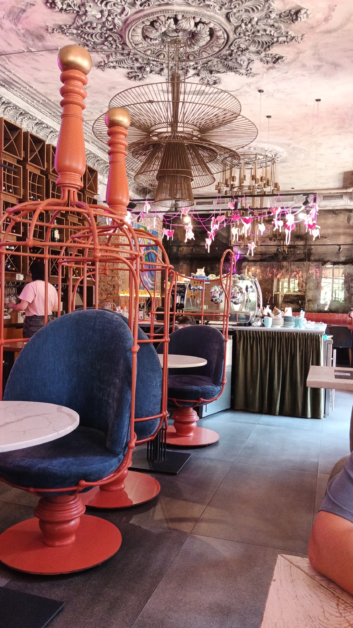 The image depicts the interior of a cafe, likely a coffee shop or restaurant, with an artistic and whimsical decor. In the foreground, there are two distinctive chairs designed to resemble birdcages, with an orange metal frame and deep blue upholstered seats. These chairs are placed around circular tables with white tops.
The ceiling is ornately decorated with plaster moldings and features large, modern chandeliers. A string of pink heart-shaped decorations hangs across the ceiling, contributing to the overall charming aesthetic.
In the background, there is a bar area with a coffee machine and shelves filled with bottles. A woman wearing a pink shirt is visible behind the bar, appearing to be working. The walls have exposed brick sections, and a large mirror reflects the interior. The flooring is made of large, dark gray tiles.
The scene appears to be well-lit, with a mix of ambient light and decorative lighting from the chandeliers and heart decorations. The overall impression is of a cozy and stylish establishment.