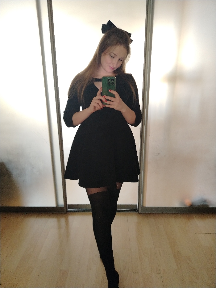 A single female individual is depicted in an indoor setting, identified as Chaikovsky, Russia. She stands centered, facing a reflective surface and holding a smartphone with both hands, appearing to take a mirror selfie. Her head is tilted downwards, and her gaze is directed towards the phone screen. She has long, light brown hair styled with a black bow at the crown. She is dressed in a solid black, knee-length dress featuring a V-neckline with a horizontal cutout, three-quarter sleeves, and a flared skirt. She is also wearing black over-the-knee stockings. The smartphone held in her hands has a green protective case, and her fingernails are painted red. The background consists of two large, vertical, rectangular panels with a frosted or translucent finish, likely sliding doors of a wardrobe or closet, showing faint, blurred reflections. The floor is covered with light-toned wooden planks arranged horizontally.