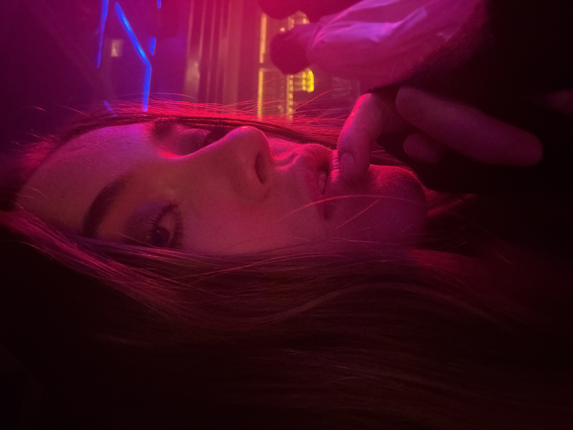 The image displays an indoor scene under predominant red and pink neon lighting, consistent with a nightclub, bar, or lounge environment in Mexico City, Mexico. A female subject occupies the foreground, illuminated by intense red and pink light sources. Her left eye is visible, directed towards the viewer, while her right index finger touches her lips. Her long, dark hair is prominent. In the blurred background, to the right, the silhouette of another individual wearing a light-colored top is discernible. Behind this figure, shelves stocked with bottles are visible as part of a bar area, illuminated with amber light. Overhead, blue geometric neon light fixtures are present. The specific venue within Mexico City cannot be determined from the visual evidence.