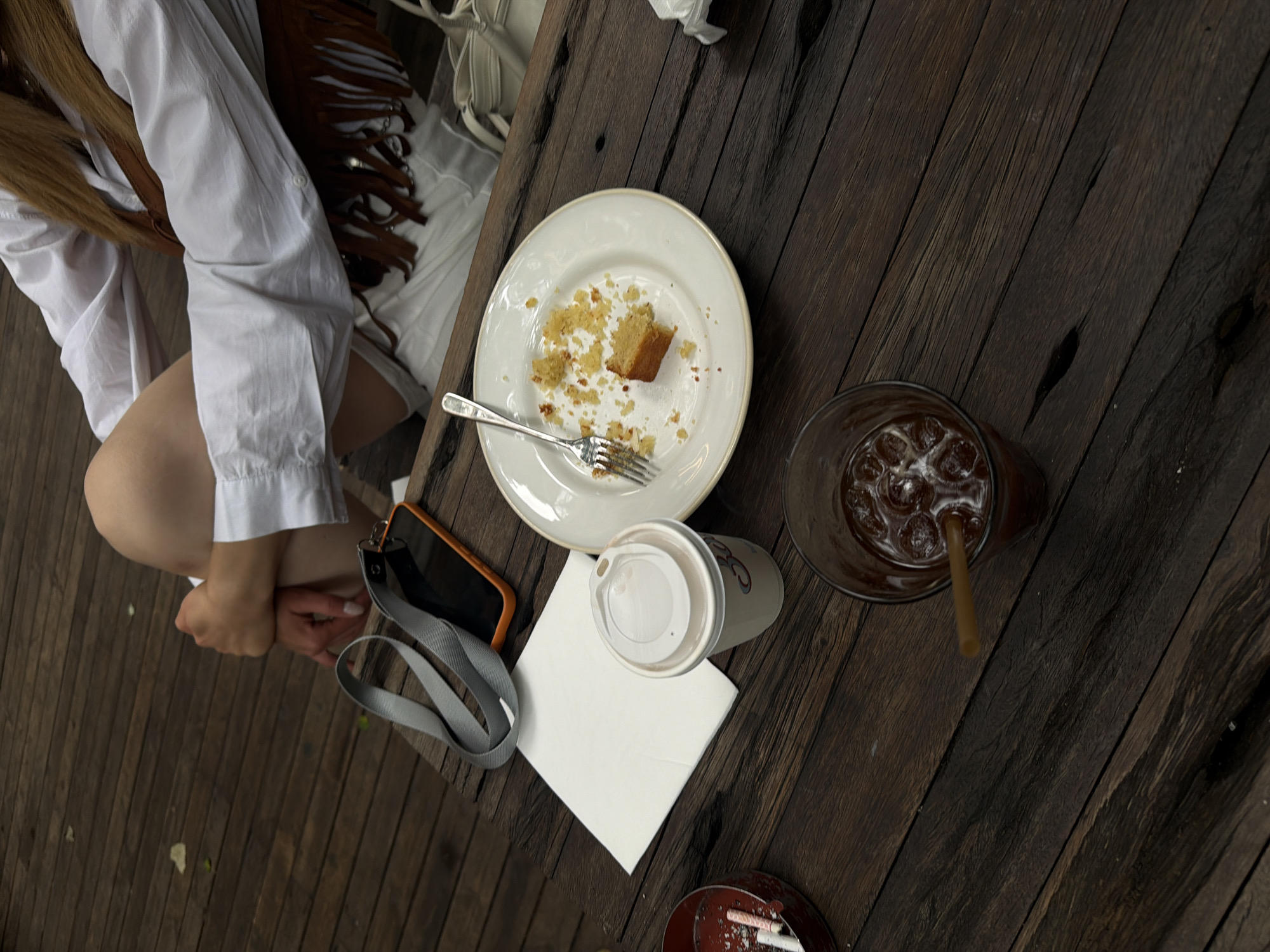 An outdoor scene on a wooden deck, likely in Tbilisi, Georgia, shows a person sitting with their legs drawn up. In the foreground, a dark wooden table holds several objects: a glass with ice and a brown beverage with a straw, a white paper cup with a lid, a folded white napkin, a plate with a piece of cake and scattered crumbs, a fork, and an orange-cased smartphone with a grey lanyard. To the left, a small round ashtray contains several cigarette butts. The person is wearing a white shirt and a brown fringed vest. The lighting suggests it is daytime.