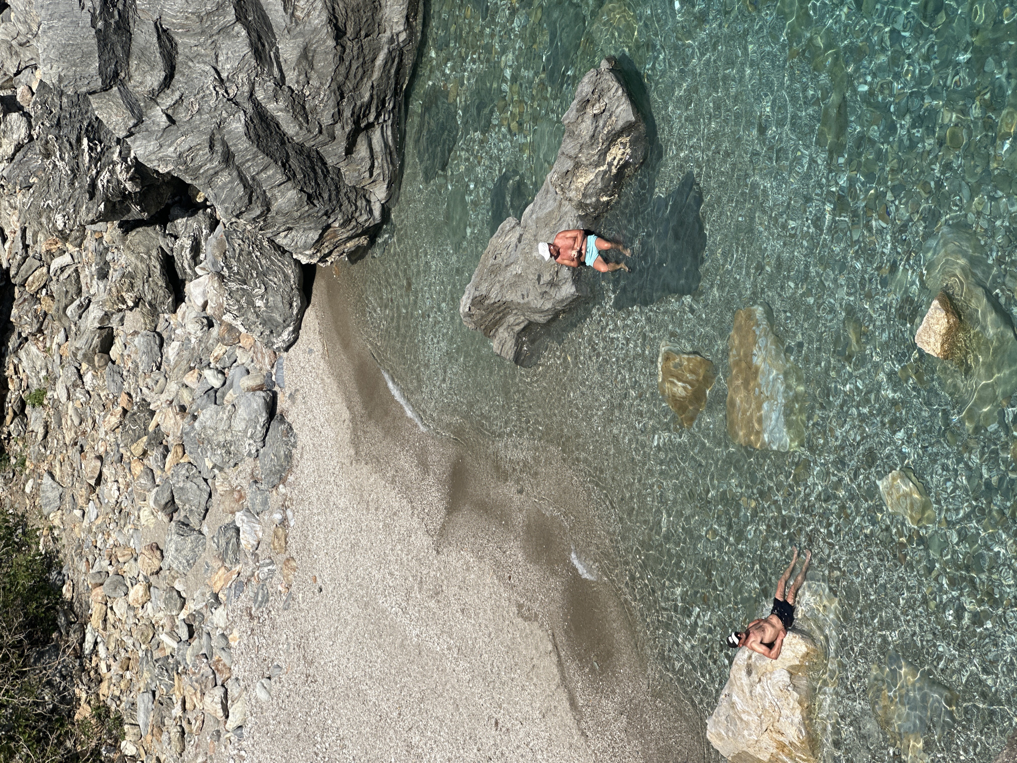 The image shows a section of a rocky beach and clear water, presumably near Gazipaşa, Turkey. Large grey rocks and smaller pebbles form the beach, with a small wave visible where the water meets the sand. Two people are in the water. One is lying on a large rock wearing a white hat and black swim trunks. The other, also wearing a white hat and light blue swim trunks, is floating on their back next to another large rock. The water is clear enough to see the pebbles and rocks beneath the surface.