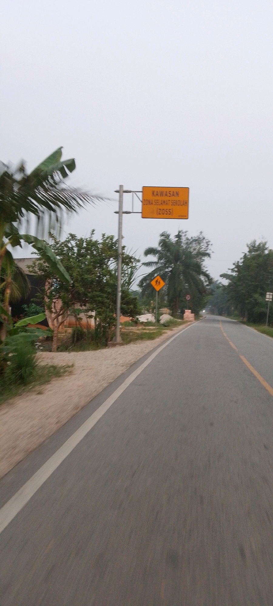 The image shows a road in Lenggadai Hulu, Indonesia. On the left side of the road, there are trees and a house. There is also a signpost with a yellow sign that says "KAWASAN ZONA SELAMAT SEKOLAH (ZOSS)". Further down the road, there is a yellow diamond-shaped sign with a black silhouette of two children walking. The road has a white line on the left and a yellow line on the right. The sky is overcast.