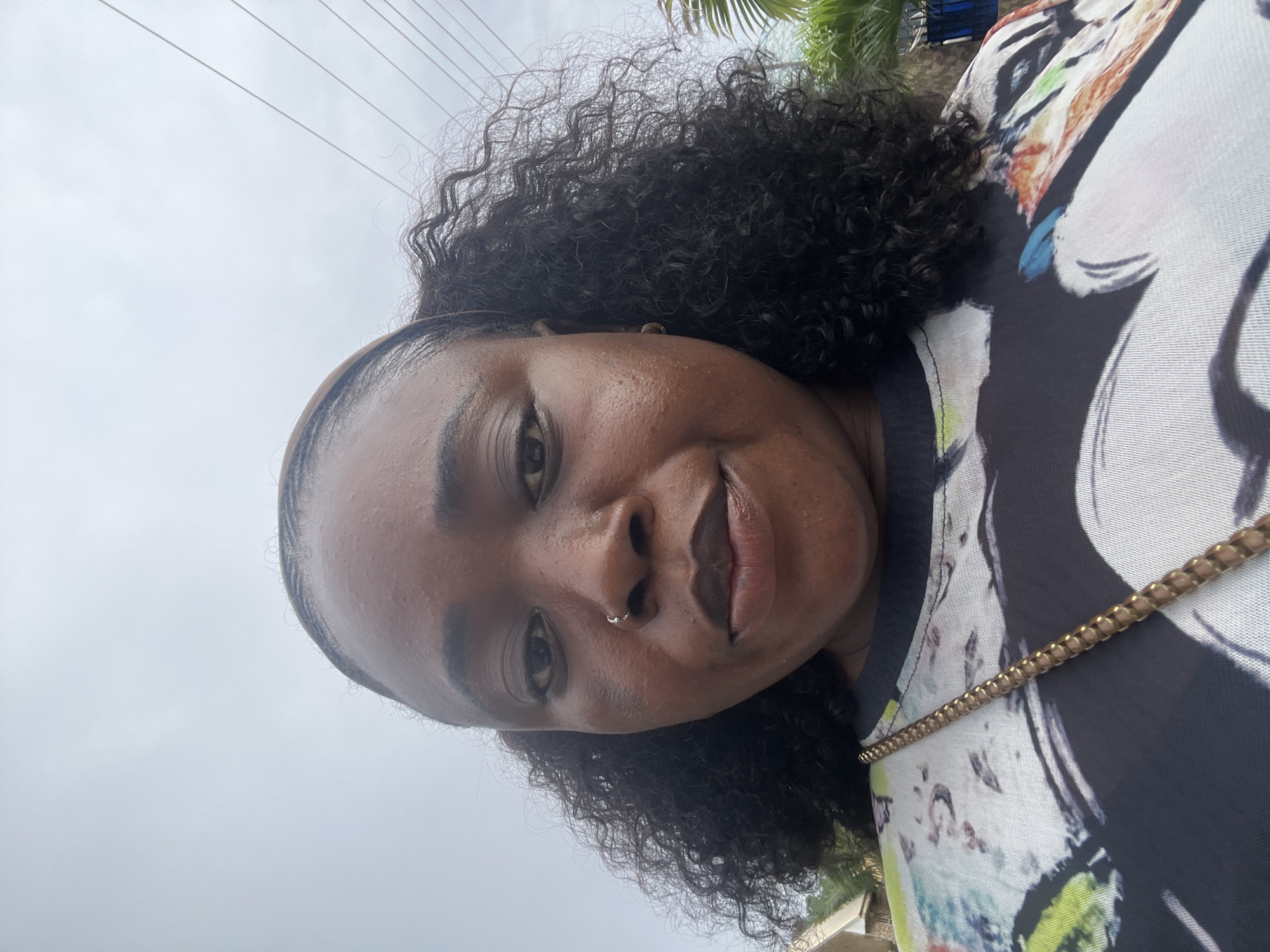 This is a close-up, medium-angle, outdoor selfie of a Black woman taken from a low perspective against a cloudy sky. The woman is facing the camera, with her head tilted slightly downwards. She has dark, curly hair, a visible hairband, and a nose ring. Her skin has a slight sheen. She is wearing a dark t-shirt with a colorful, abstract design featuring bold strokes of black, white, green, and orange. A thin, gold chain strap of a purse is visible crossing her chest over the t-shirt. In the background, slightly out of focus, are some trees and what appears to be part of a building. Utility wires are visible in the upper right quadrant of the image. The overall lighting suggests an overcast day. The provided context indicates the location is the city of Abuja, Nigeria.