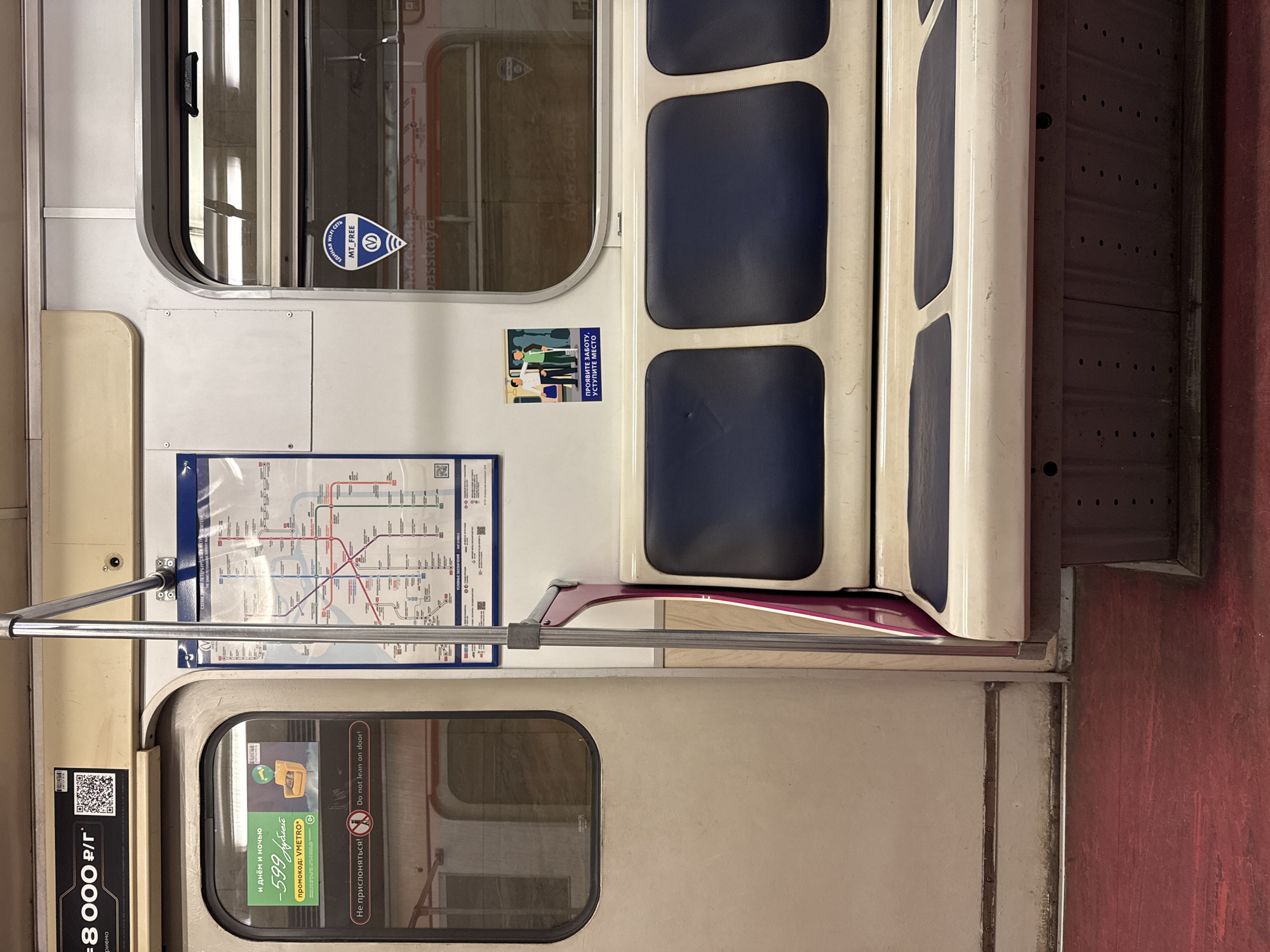 The image captures the interior of a metro car, rotated 90 degrees clockwise. The setting is a brightly lit, modern train cabin, likely in Saint Petersburg, Russia, as suggested by the visible station name "passkaya" (referring to Spasskaya station) and Russian text.
On the left side of the frame, a window is visible, reflecting the interior and some text. Below it, a white panel precedes a transparent holder displaying a metro map, which shows various colored lines and station names. A silver horizontal grab bar runs across the mid-section of the image.
The central part of the image features a row of blue-cushioned seats with white frames, typical of public transport. To the right of the seats, a dark red, textured surface forms either a lower wall panel or part of the floor.
Several informational and advertising stickers are present. On the upper window, a circular blue sticker indicates "ЕДИНАЯ WI-FI СЕТЬ MT_FREE M" (Unified Wi-Fi Network MT_FREE M), alongside a distinctive metro logo. Below the seats, a sticker depicts people and reads "ПРОЯВИТЕ ЗАБОТУ, УСТУПИТЕ МЕСТО" (Show care, give up your seat), promoting courtesy. On the door's window, a green sticker advertises "и днём и ночью -599 рублей промокод: VMETRO" (day and night -599 rubles promo code: VMETRO), accompanied by a QR code. Another sticker next to it provides a warning in both Russian and English: "Не прислоняться!" and "Do not lean on door!". Further to the left, near the metro map, a partially visible QR code and text "=8000 р/Г" are present. No people are visible, and the scene suggests a quiet moment inside the train car.