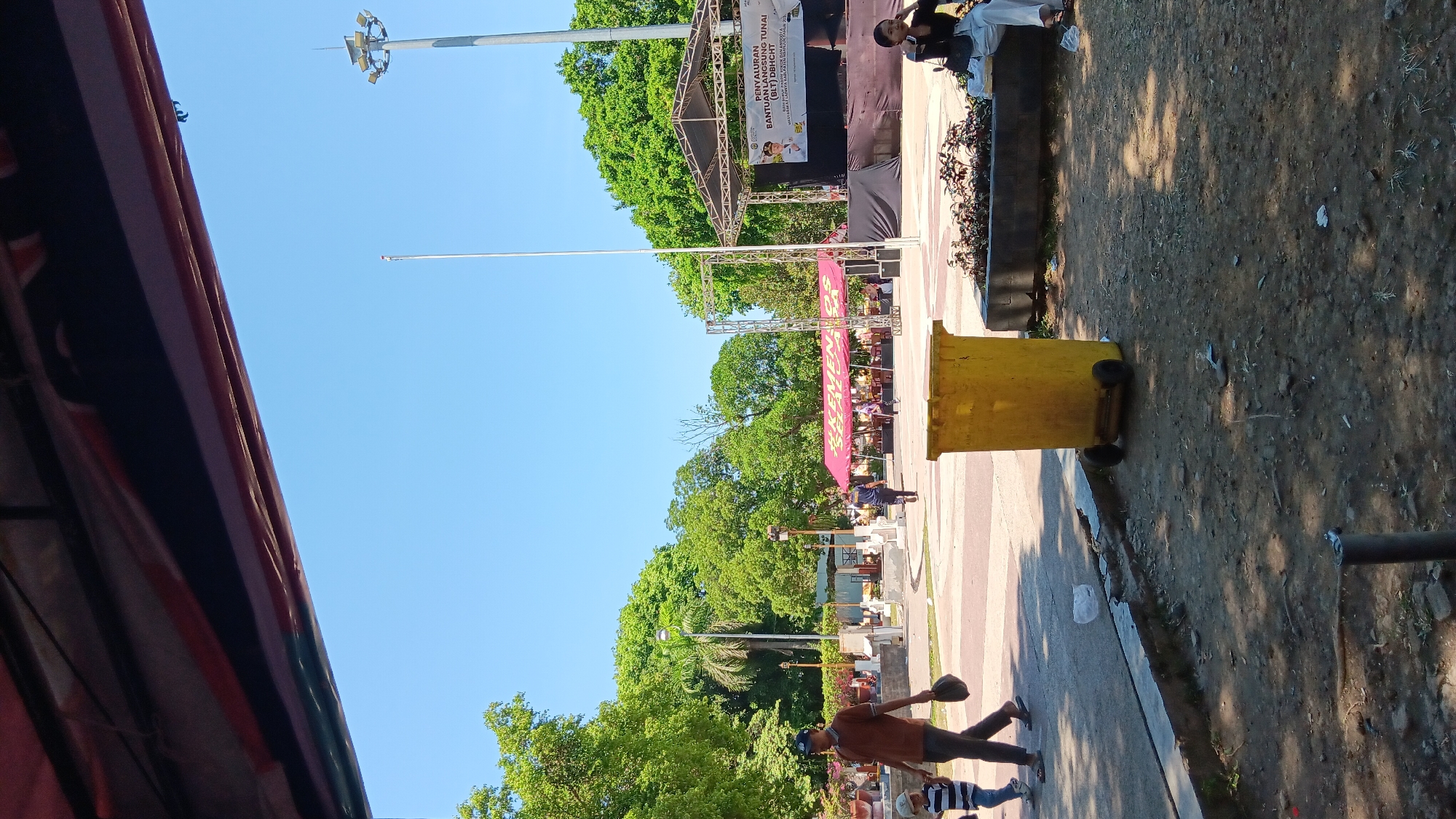 The image depicts an outdoor scene in a public park, likely in Nganjuk, Indonesia, under a clear blue sky. A stage structure with a banner is visible on the right side, suggesting an event is taking place or has recently concluded. The banner displays text that appears to be in Indonesian, possibly related to aid distribution. A bright yellow trash can with wheels is positioned prominently in the foreground. Several people are scattered throughout the park: a man and a child are walking on a paved area on the left, a person in a dark uniform is walking towards the right, and another person is seated on a bench on the far right. Lush green trees surround the park, with a tall light pole visible in the background. The ground is a mix of paved surfaces and dirt. A red tent is also present to the left of the stage.