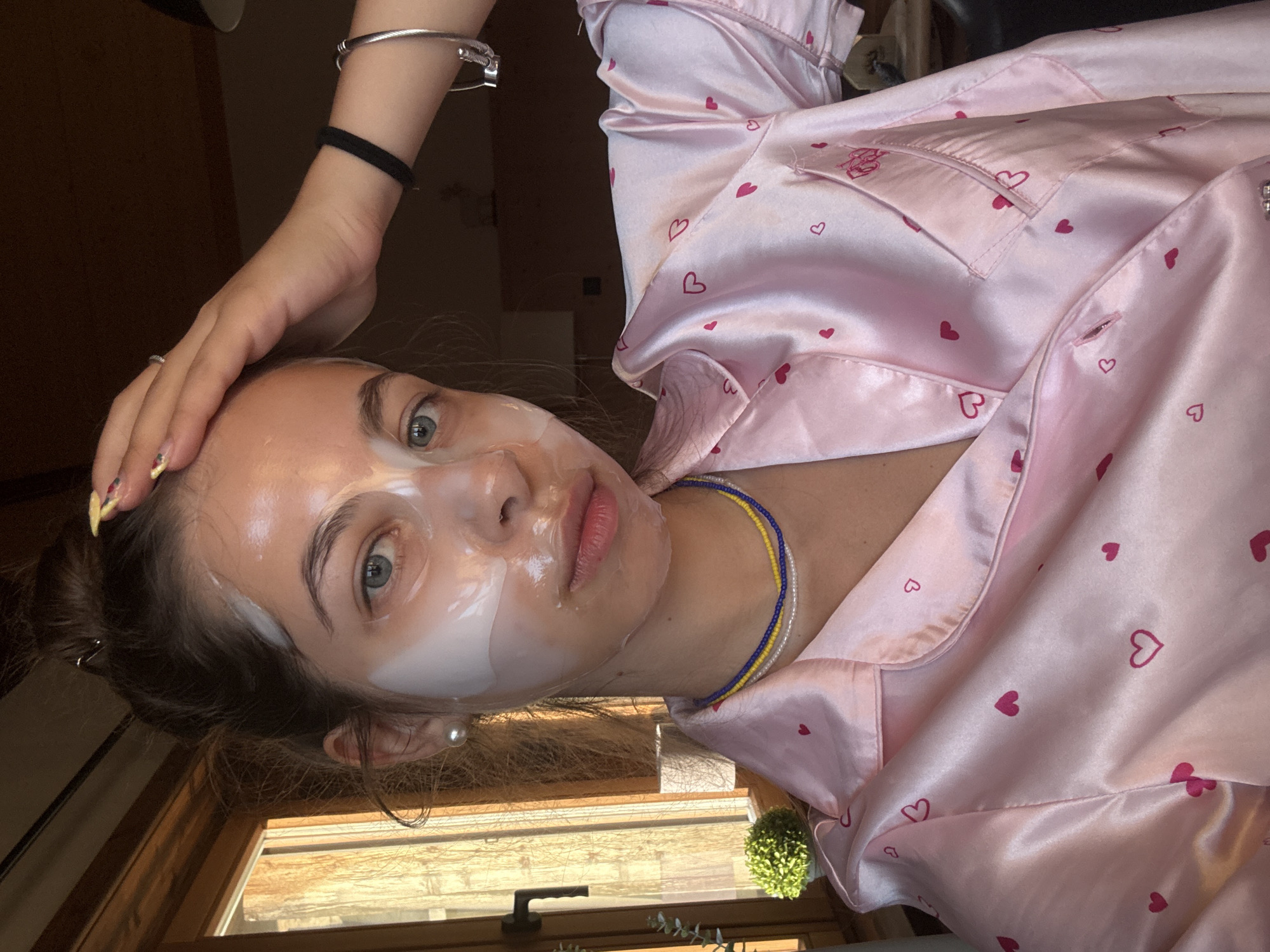 A young woman is shown from the chest up, wearing a pink satin pajama top adorned with small red hearts. She has a sheet mask on her face, covering her eyes and nose, with portions of it peeling up around the edges. Her hair is pulled up in a messy bun, and a few strands fall around her face. She has light blue eyes and is looking directly at the camera. She is wearing a pearl earring in her left ear, and a multi-colored beaded necklace around her neck. Her right hand is resting on her forehead, and her left arm is visible, adorned with a black hair tie and a silver bracelet.
The background appears to be indoors, with a window to the left showing a sunlit outdoor scene. The window frame is made of wood, and a small potted plant is visible on the windowsill. The lighting suggests it is daytime. The scene does not provide any specific indicators to place it in Breungeshain, Germany.