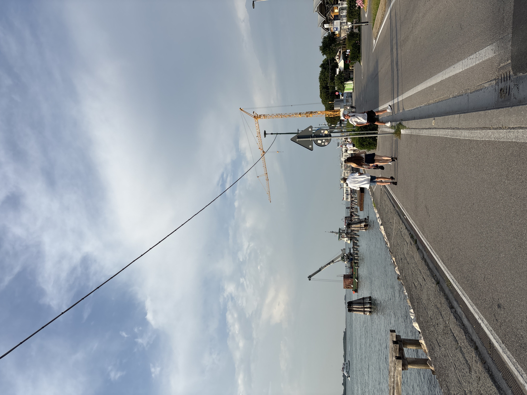 The scene is an outdoor view in Punta Sabbioni, Italy. A long electrical wire stretches diagonally across the sky. In the background are boats in the water near the coast. A yellow crane stands on the coast near the boats. There are people walking on the sidewalk near the coastal road, including a man in a white shirt and dark shorts and two other people walking alongside.