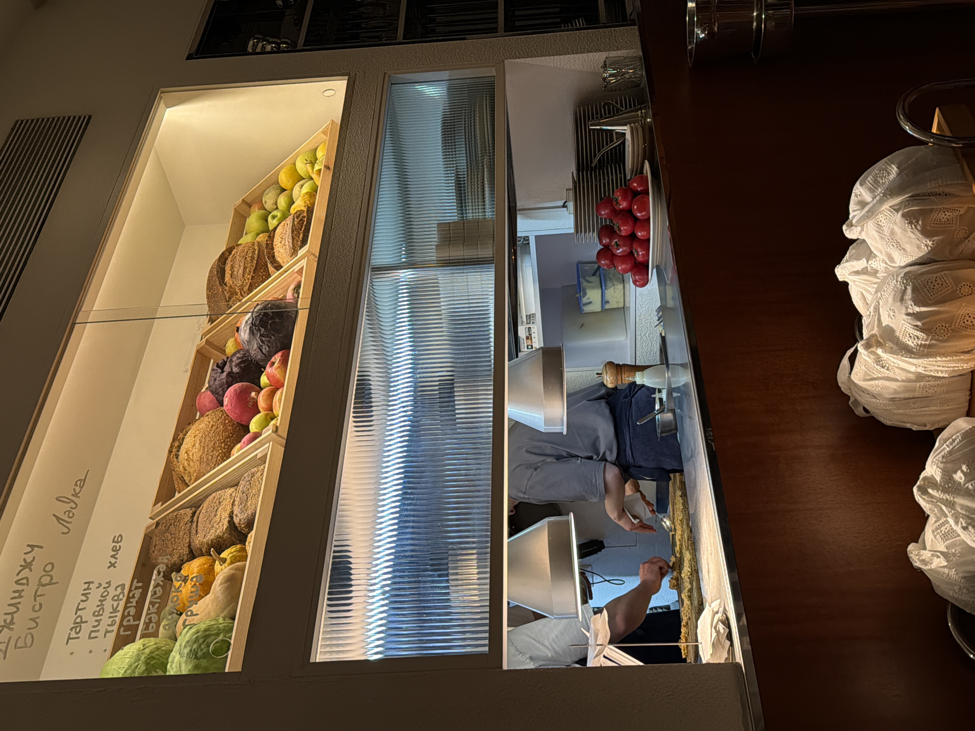 The image shows a view of a deli or bakery counter in Moscow, Russia. The left section displays a variety of fresh produce and baked goods, including apples, pears, bread, cabbage, and pumpkins, arranged on shelves with Cyrillic text visible. In the center is a panel of textured glass. The right section provides a glimpse into the kitchen, with employees preparing food, including a plate of tomatoes. Stacks of white fabric, possibly napkins or dishcloths, are seen on the counter.