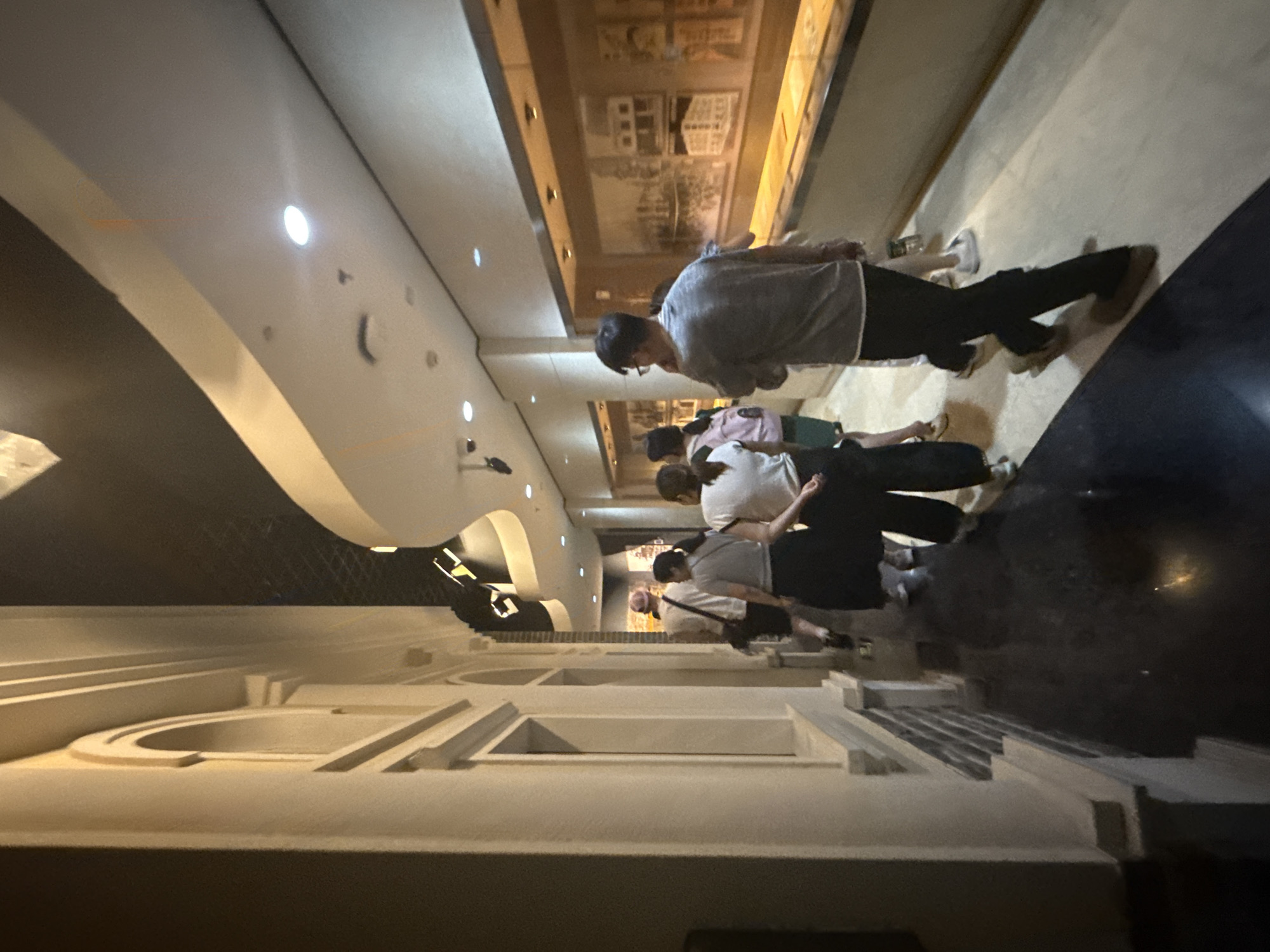 A group of people are walking down a museum hallway in Yuzhong District, China. The hallway is dimly lit, with architectural elements on the left side resembling classical columns and arched windows. On the right side of the hallway, there are display cases showcasing what appear to be historical photographs or artwork. The people in the group are casually dressed and are moving in the same direction, suggesting they are touring the museum. The floor is reflective and dark, adding to the moody ambiance of the space.
