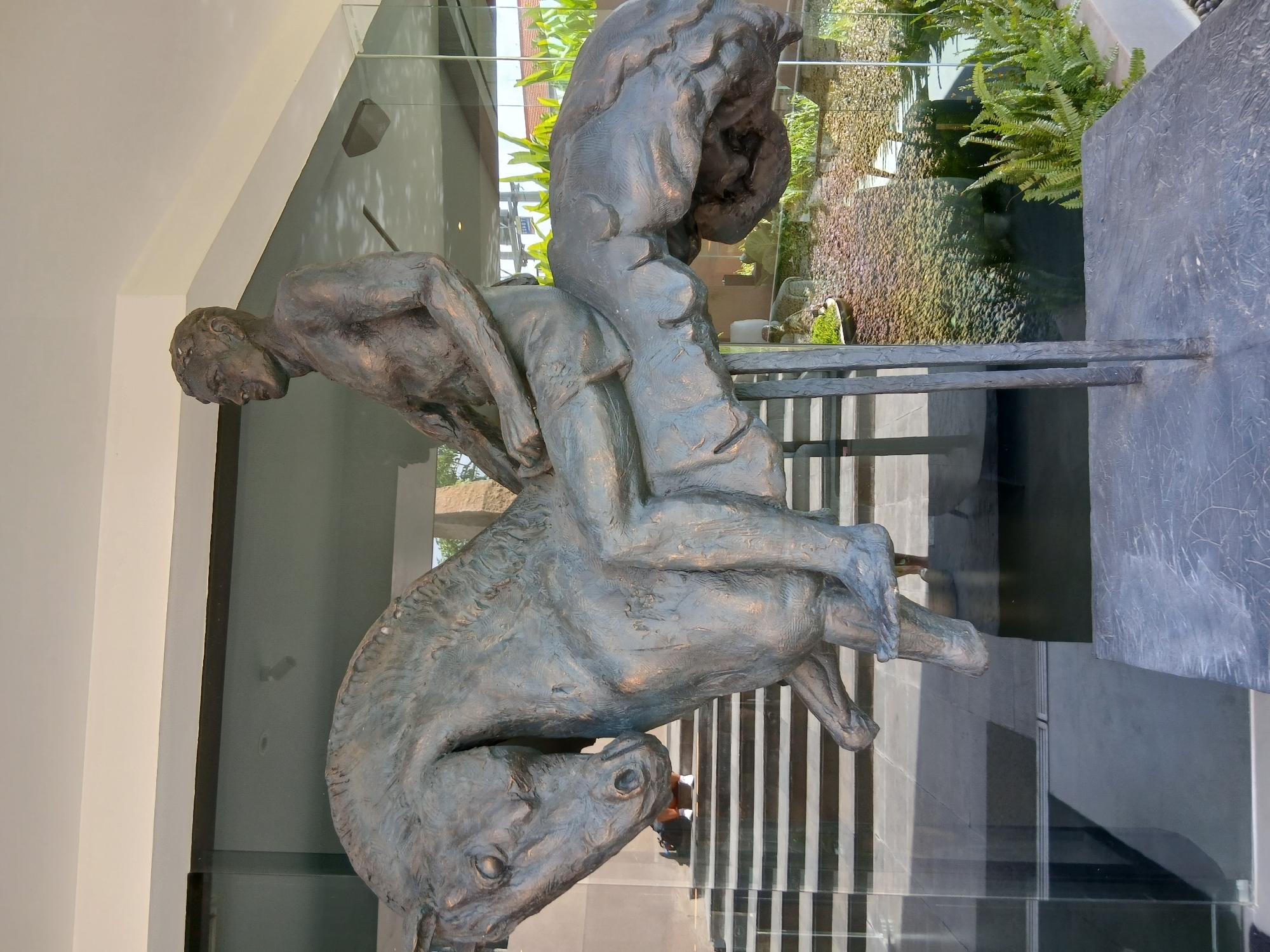 A bronze sculpture of a man riding a horse is displayed outdoors in front of a building with large glass windows. The sculpture is mounted on a dark base supported by two metal legs. The man is depicted with a muscular physique, wearing shorts. The horse is shown in mid-stride, with its head lowered. Through the glass windows, a staircase with black and white steps is visible, along with lush green foliage and what appears to be a small pond with decorative stones. The overall setting suggests an urban or commercial environment, possibly a hotel or public space, in North Kuta, Indonesia.