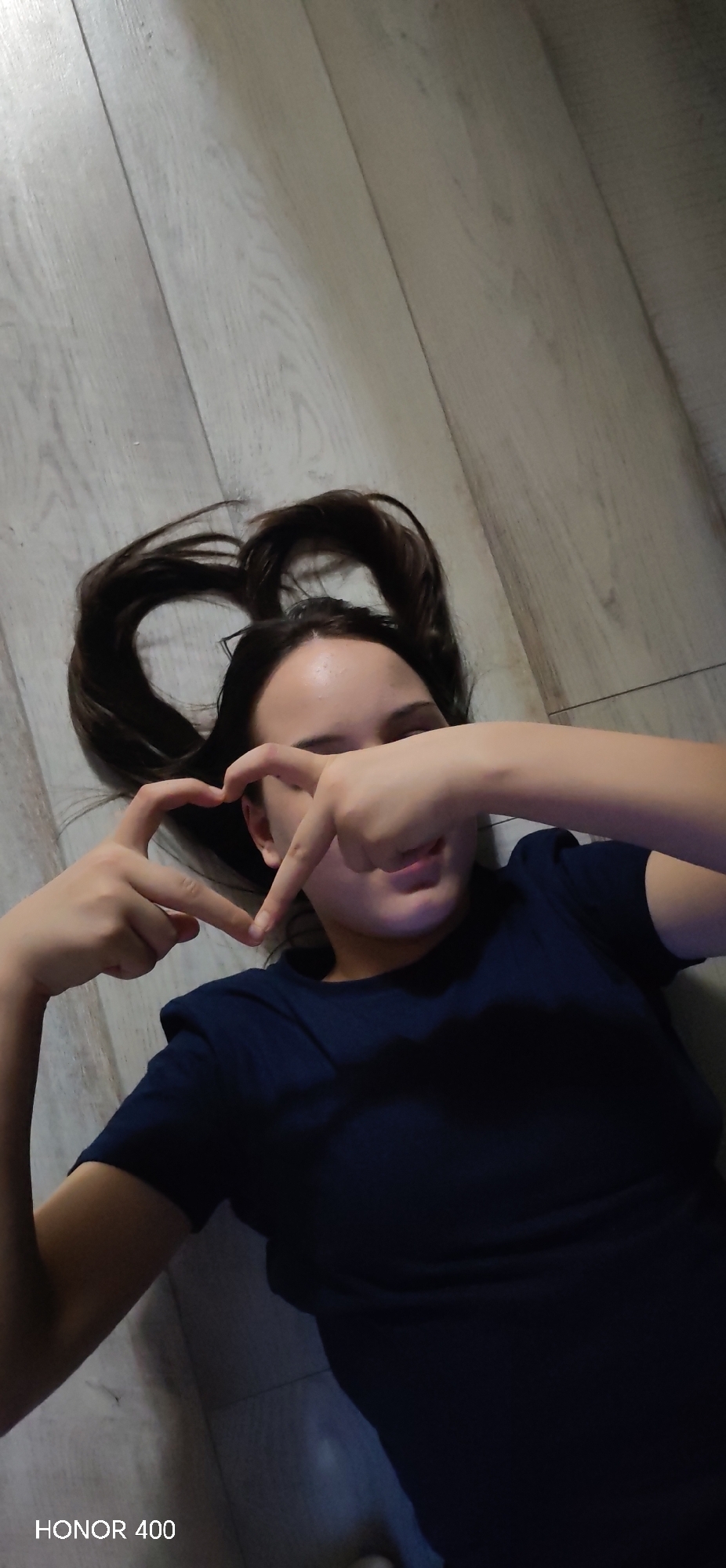 A young female is lying on a light-colored, wood-grain floor, possibly made of laminate or vinyl. Her dark hair is styled in a heart shape above her head. She is making a heart shape with her hands, positioned in front of her face. She is wearing a dark blue t-shirt. The lower left corner of the image shows text "HONOR 400". Given the model of the phone used, it's plausible the location is in Noviy, Russia.