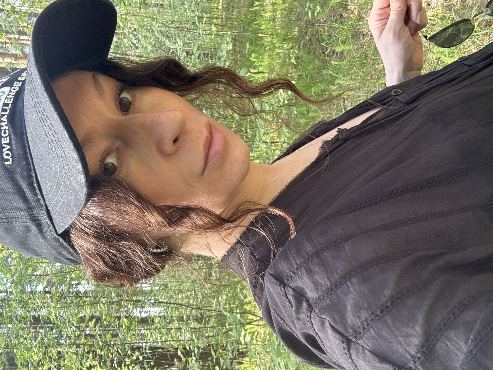 A woman wearing a black baseball cap with white lettering, a black shirt, and earrings is holding sunglasses in her right hand. She is outdoors, with greenery visible in the background, possibly in a wooded area. The image appears to have been taken in daylight. There is no verifiable evidence to confirm the location of the photograph is in Chaikovsky, Russia.