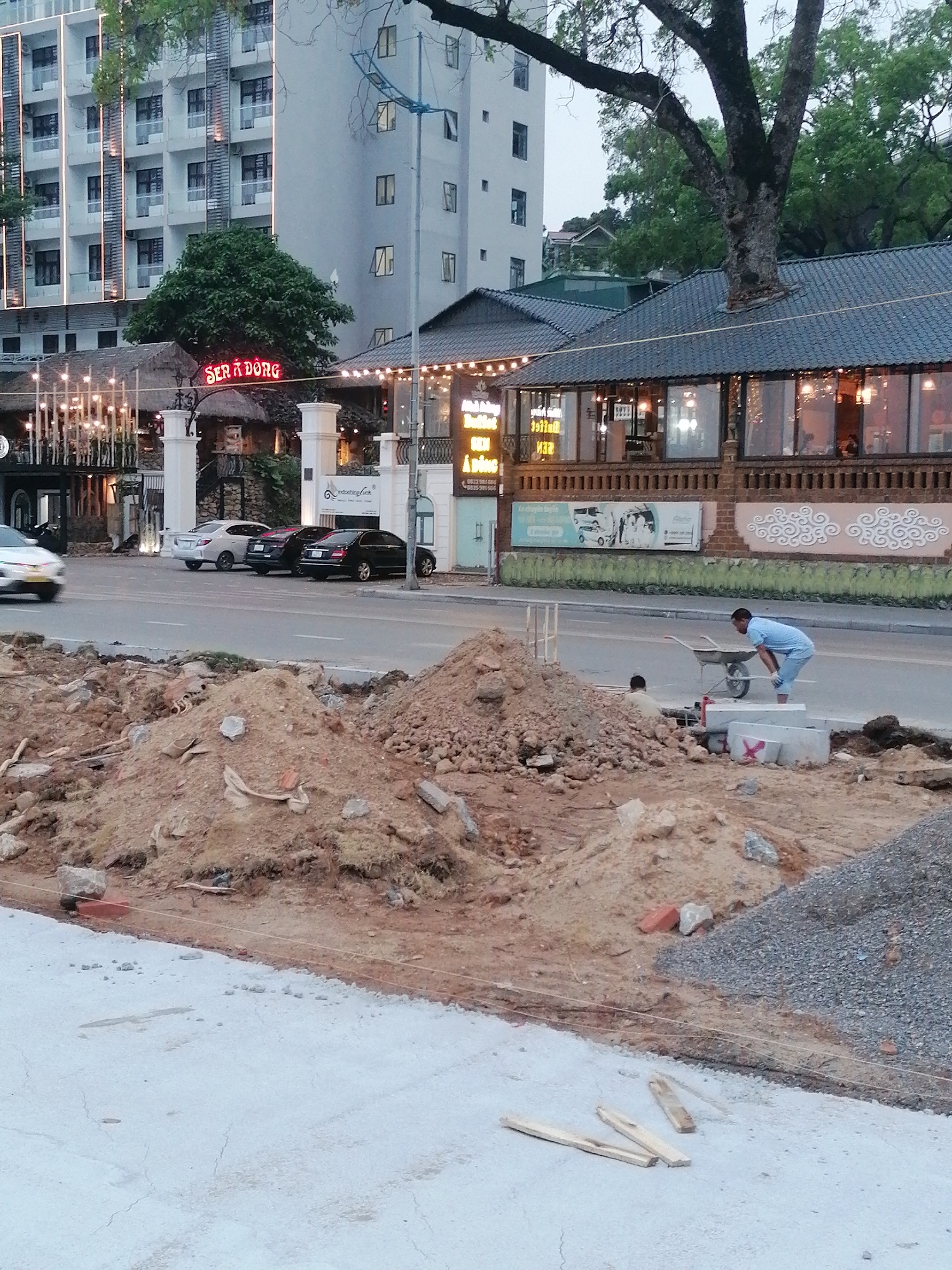 The image shows a construction site in Bai Chay Ward, Vietnam. Mounds of dirt and gravel are present on the foreground. A man in a light blue outfit is working with a wheelbarrow near concrete blocks marked with a red "X". A second person is crouched near the dirt piles. Behind the construction area is a street with cars parked and moving. A restaurant with a sign reading "SER A DONG" is visible, as well as other businesses and multi-story buildings. A large tree is present in the background.
