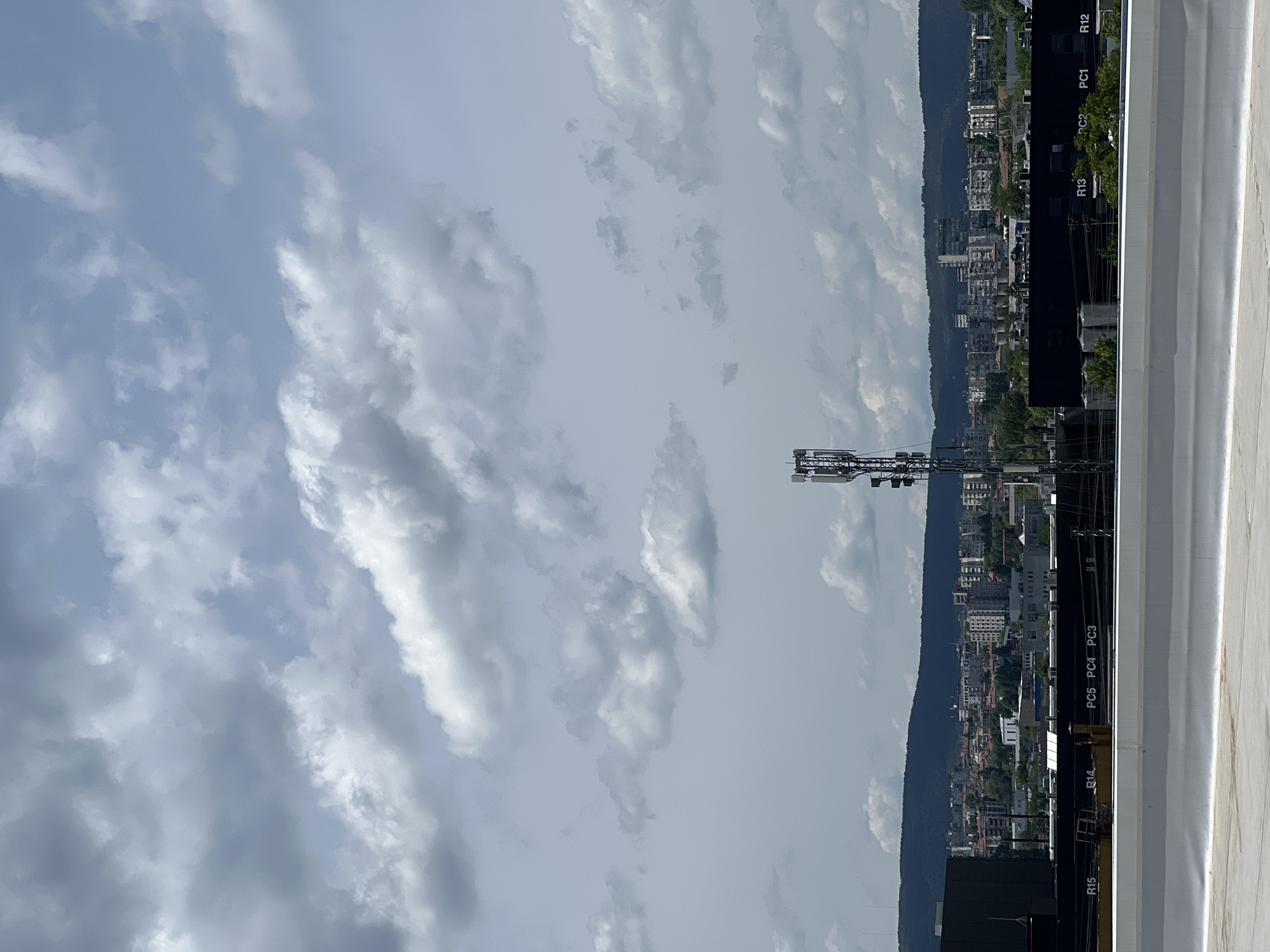 The image presents a wide view of an urban landscape set against a mountainous backdrop, seen from an elevated perspective, likely a rooftop. The sky above is largely covered with scattered, bright white and grey cumulus clouds, with visible patches of lighter blue sky, indicating daytime conditions.
In the foreground, on the right side, a flat, light-colored surface, presumably a rooftop, occupies a significant portion. Adjacent to this, a dark, linear structure runs horizontally, featuring several white alphanumeric labels. Clearly visible among these labels are "R12", "PC1", "PC2", "R13", "PC5", "PC4", "PC3", "R14", and "R15", suggesting identification markers for different sections or utilities.
The middle ground is dominated by a sprawling urban or suburban area with numerous multi-story buildings and interspersed green spaces, extending horizontally across the frame. Behind this built environment, a long, tree-covered mountain range forms a continuous, dark silhouette against the lighter sky and distant cityscape.
On the left side of the middle ground, a tall metal lattice structure stands prominently, equipped with multiple antennas and dishes, identifying it as a communication tower, possibly for telecommunications. There is no specific activity or event taking place; the image captures a static, panoramic landscape.