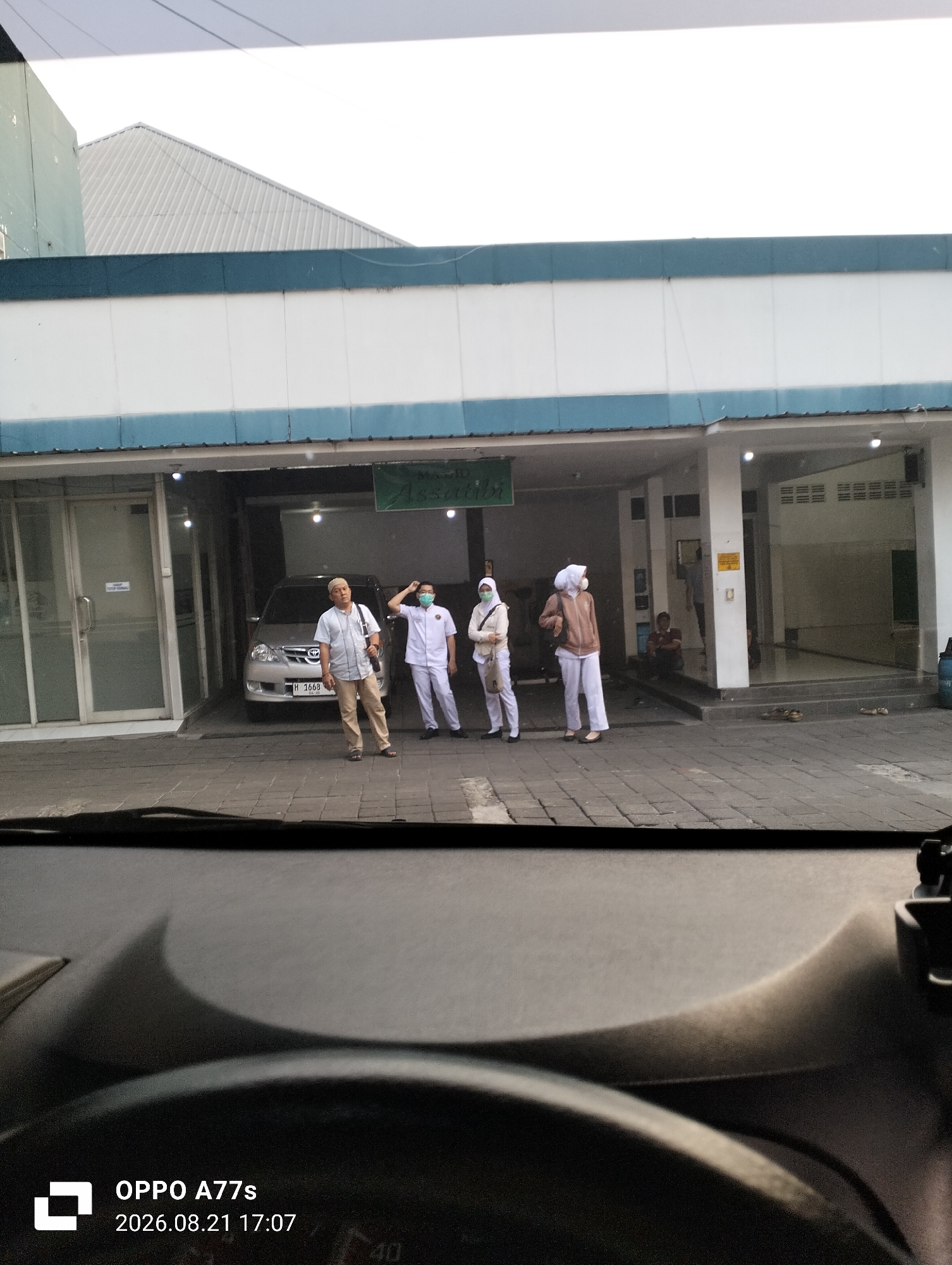 The image captures a street scene outside a building that appears to be a mosque or religious institution, based on the sign "MASJID Assalikin" visible in green lettering. Four individuals are standing on a paved area in front of the building's entrance. Two individuals on the left are wearing white uniforms and face masks, suggesting they are medical professionals or staff. One of them is a man in a light-colored shirt and khaki pants, standing with his hands by his side. The other three individuals are dressed in what appear to be uniforms or religious attire, including headscarves. To the left of the entrance is a glass-doored room with a sign inside. A silver car is parked in the driveway. The building has a white facade with a blue trim and a sloping roof. The lighting suggests it is either early morning or late afternoon. The foreground of the image shows the interior of a vehicle, likely a car, with a dashboard and the timestamp "2026.08.21 17:07" overlaid, indicating the time and date the photo was taken. The context of Semarang, Indonesia, is provided as the location.