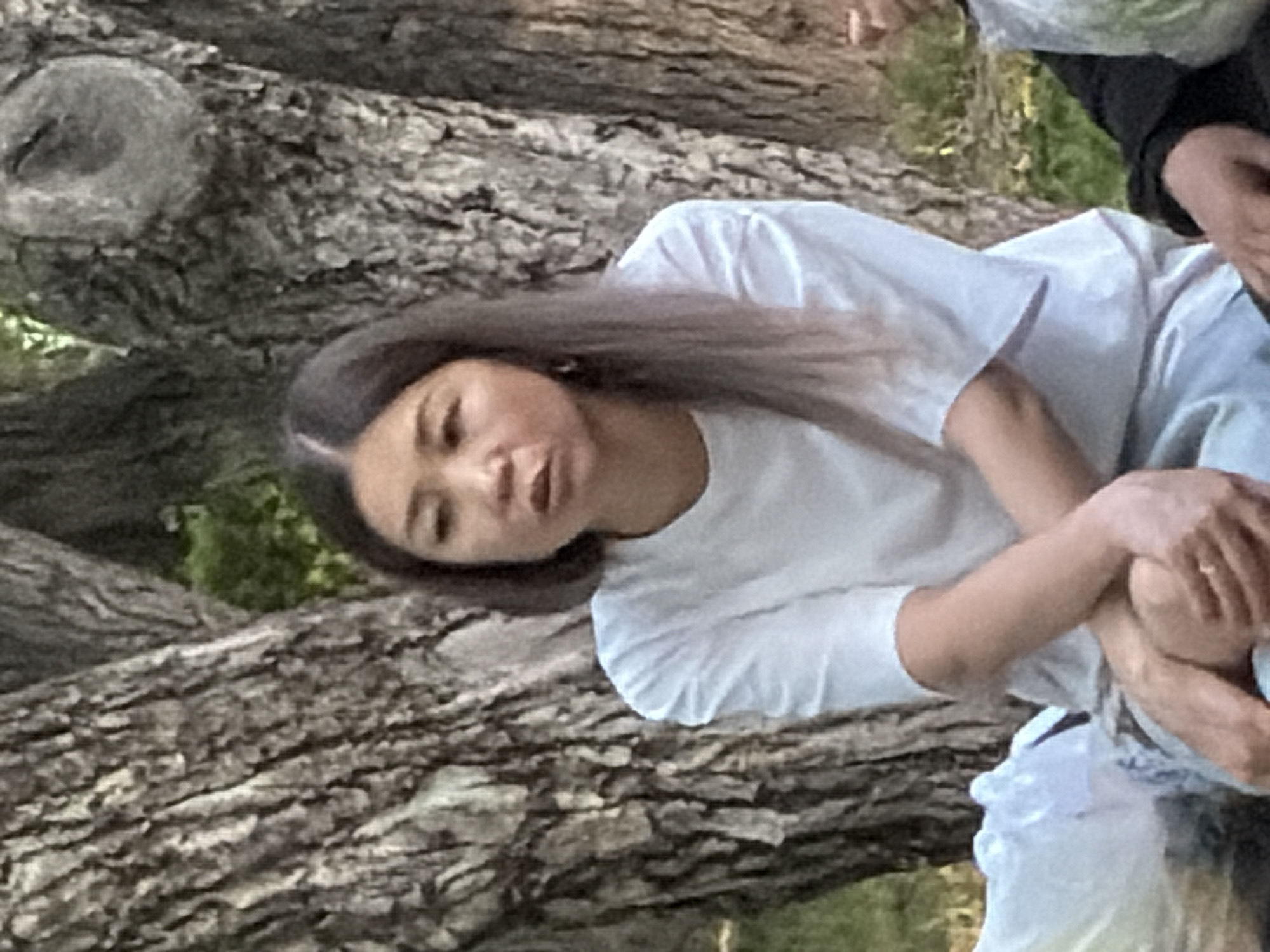 An East Asian woman with shoulder-length dark hair, some strands of which have a light purple tint, is seated in front of two large trees. She is wearing a plain white t-shirt and light blue pants. Her arms are crossed, and her hands are clasped. She has a somewhat concerned expression on her face. The background consists of the textured bark of the trees and green foliage. To the left of the woman, a white plastic bag is visible. To the right, another person in a black shirt is partially visible, also with a white plastic bag. The scene appears to be outdoors in a park or wooded area.