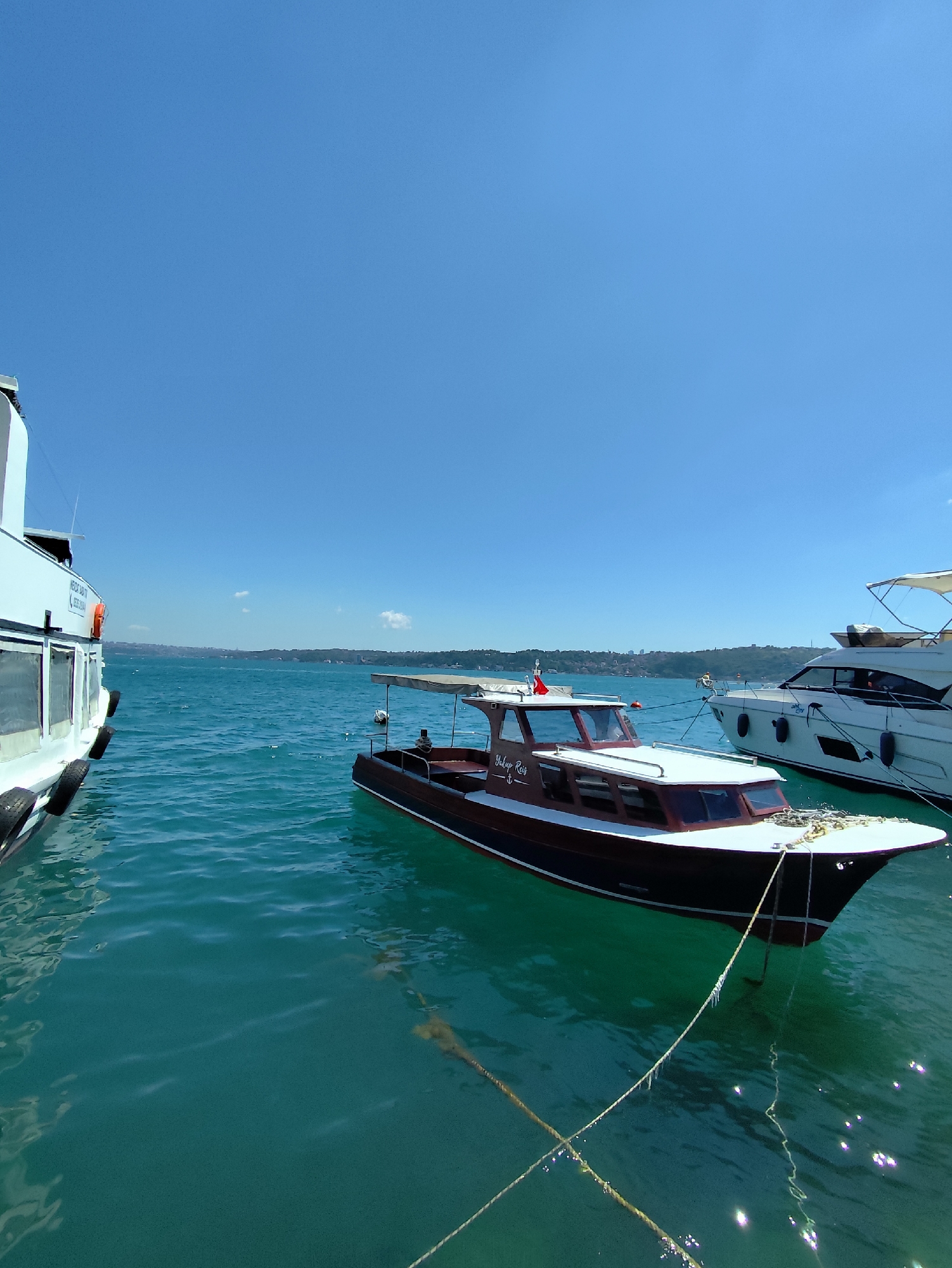 A wide-angle, eye-level shot captures a bright, sunny day on a body of water with several boats docked or moored. In the foreground, a dark-hulled boat with a white roof and a tan awning is tethered to the shore. The name "Yalup Reis" is visible on its side. To the left, the stern of a larger white boat, possibly a ferry or cruise ship, is partially visible. To the right, a sleek white yacht with a white awning and dark windows is also moored. The water is a vibrant turquoise and appears calm, with gentle ripples reflecting the sunlight. In the background, a distant, hazy coastline with green hills and some structures can be seen under a clear blue sky. The scene suggests a leisurely day at a marina or harbor.