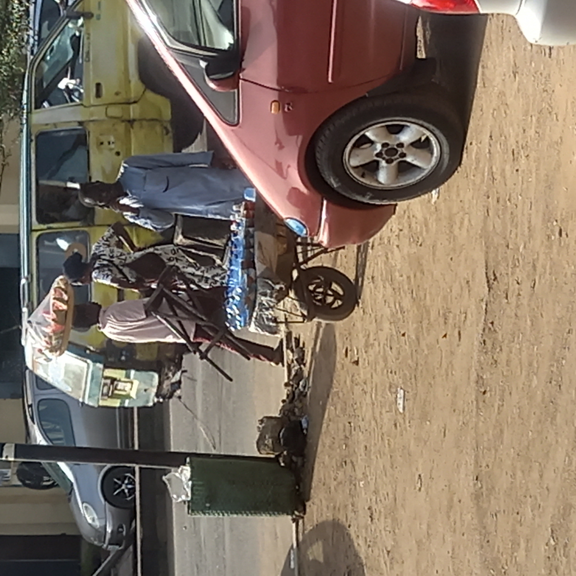 This image captures a vibrant, sunlit street scene in what appears to be Dadi Riba, Nigeria. The setting features an unpaved, dusty road with several vehicles and people engaged in daily activities.
Several individuals are visible. One person in a light blue traditional garment stands near a yellow, older model minibus or van, which appears weathered and possibly used for public transport. Another person in a patterned top is also near the yellow van, and further right, a person in a light-colored top stands beside a two-wheeled cart loaded with numerous bottles of water, indicating they are a street vendor. A fourth individual, wearing a patterned head covering and also appearing to carry something on their head, is partially visible near the vendor.
Prominently featured are multiple vehicles. A red sedan is parked or stopped close to the yellow minibus, with its passenger side and front wheel clearly visible. Its paint has some wear. The yellow minibus itself shows signs of age and possibly missing parts, hinting at its functional, utilitarian role. In the background, further down the road, a grey car is partially visible.
The ground is largely dirt or sand, characteristic of unpaved roads in many regions. There's a green, possibly metal, bin or stand near the foreground, with some debris scattered on the ground around it. The bright, clear lighting suggests it is daytime, likely mid-morning or afternoon, with no discernible harsh shadows. The overall scene depicts a bustling, ordinary moment of street life and commerce, typical of a local community. No distinct text is legible within the image.