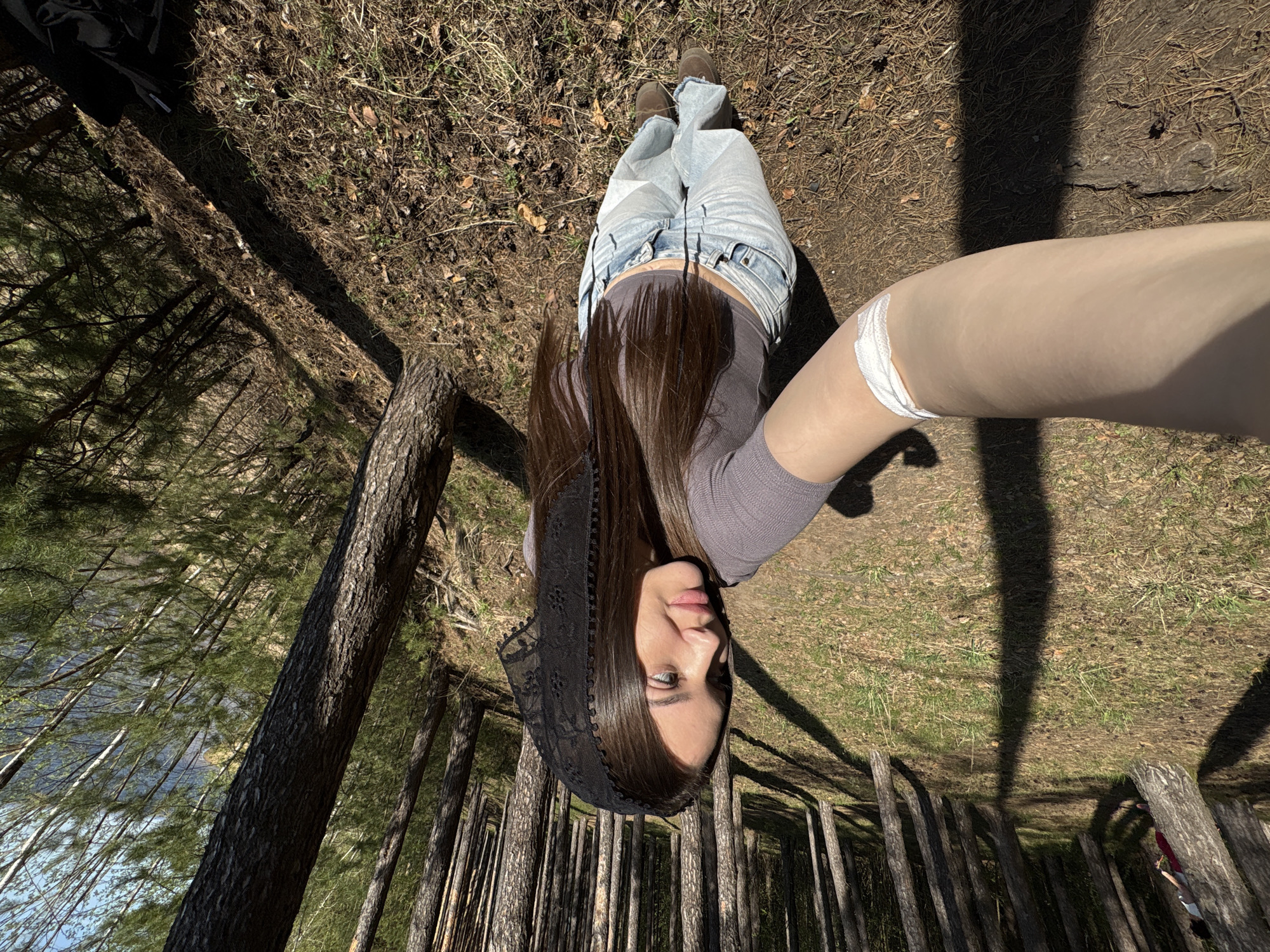 A woman is lying on the ground in a forest, taking a selfie. She wears jeans, a brown top, brown boots, and a black lace head covering. The location is a pine forest, characterized by the presence of tall, thin trees. The environment consists of dry grass, fallen leaves, and tree shadows. The city of Unknown, Unknown is not directly identifiable from the image.