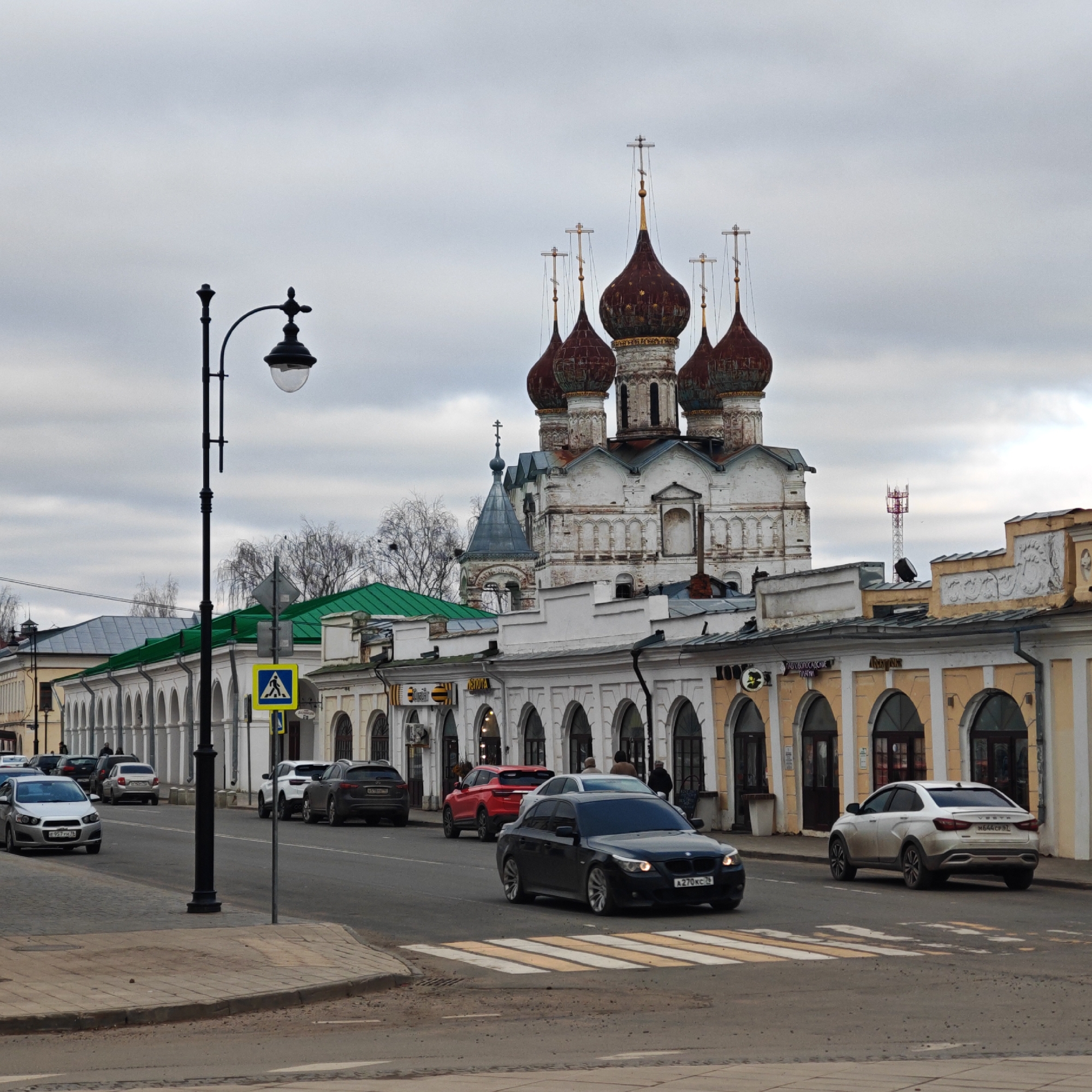 The image depicts a street scene in Rostov, Russia, under an overcast sky. Dominating the background is a large white building with several red onion domes topped with golden crosses, indicative of a Russian Orthodox church. The street is lined with buildings, many with arched facades. Various vehicles are visible on the road, including cars of different makes and colors, some stationary and some in motion. A pedestrian crossing is marked on the road, suggesting a crossing point for pedestrians. A black lamppost stands on the sidewalk on the left side of the image. Yellow pedestrian sign is visible next to the lamppost.