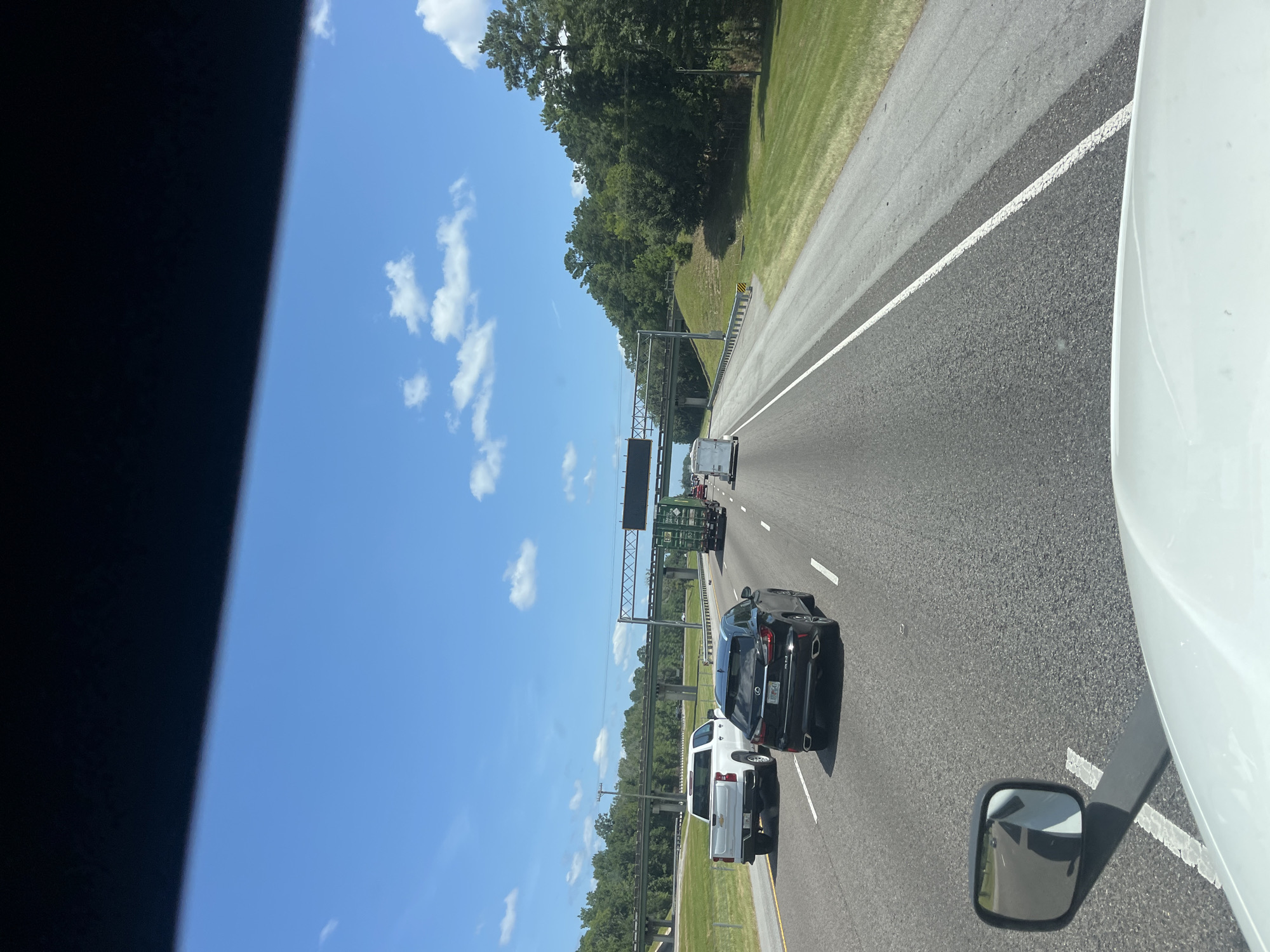 The image shows a view from the driver's seat of a vehicle on a highway, with the hood of the vehicle visible in the foreground. The road is asphalt with white lane markings. Several vehicles are traveling on the highway, including a white pickup truck, a black Lexus SUV, a green truck with a container, and a white trailer. Trees and vegetation line the side of the highway, and an overpass is visible in the distance. The sky is blue with scattered white clouds. The location is Riceboro, United States.