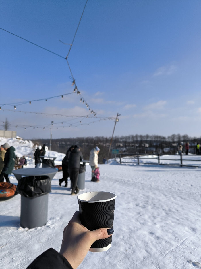A forensic examination of the image depicts an outdoor, snow-covered recreational area in Prybuzke, Ukraine, under a clear, bright blue sky with minimal cloud cover.
In the immediate foreground, a fair-skinned right hand, adorned with dark blue nail polish, holds a black, corrugated disposable cup with a white interior and rim. A portion of a black jacket sleeve is visible at the bottom left.
The midground features a pristine expanse of snow. Several individuals, blurred due to depth of field, are present, dressed in winter attire. To the left, a person in a dark green coat walks past a gray cylindrical table topped with a black trash bag. Nearby, another identical table and trash receptacle are visible. Multiple blurred figures congregate around these tables. A person in a light-colored jacket stands with a child in a pink outfit adjacent to them, both facing away from the viewer. In the upper midground, strings of illuminated decorative lights are suspended between support poles, creating an overhead canopy.
The background reveals a distant treeline of bare, dark trees stretching horizontally across the horizon. Below this, a gentle slope of snow leads to a blurred, distant landscape. To the right, a low, light-colored wooden fence runs parallel to the horizon. Additional blurred figures are visible in the far background, possibly engaging in activities on the snowy terrain. The overall scene suggests a sunny winter day at a public or recreational facility.