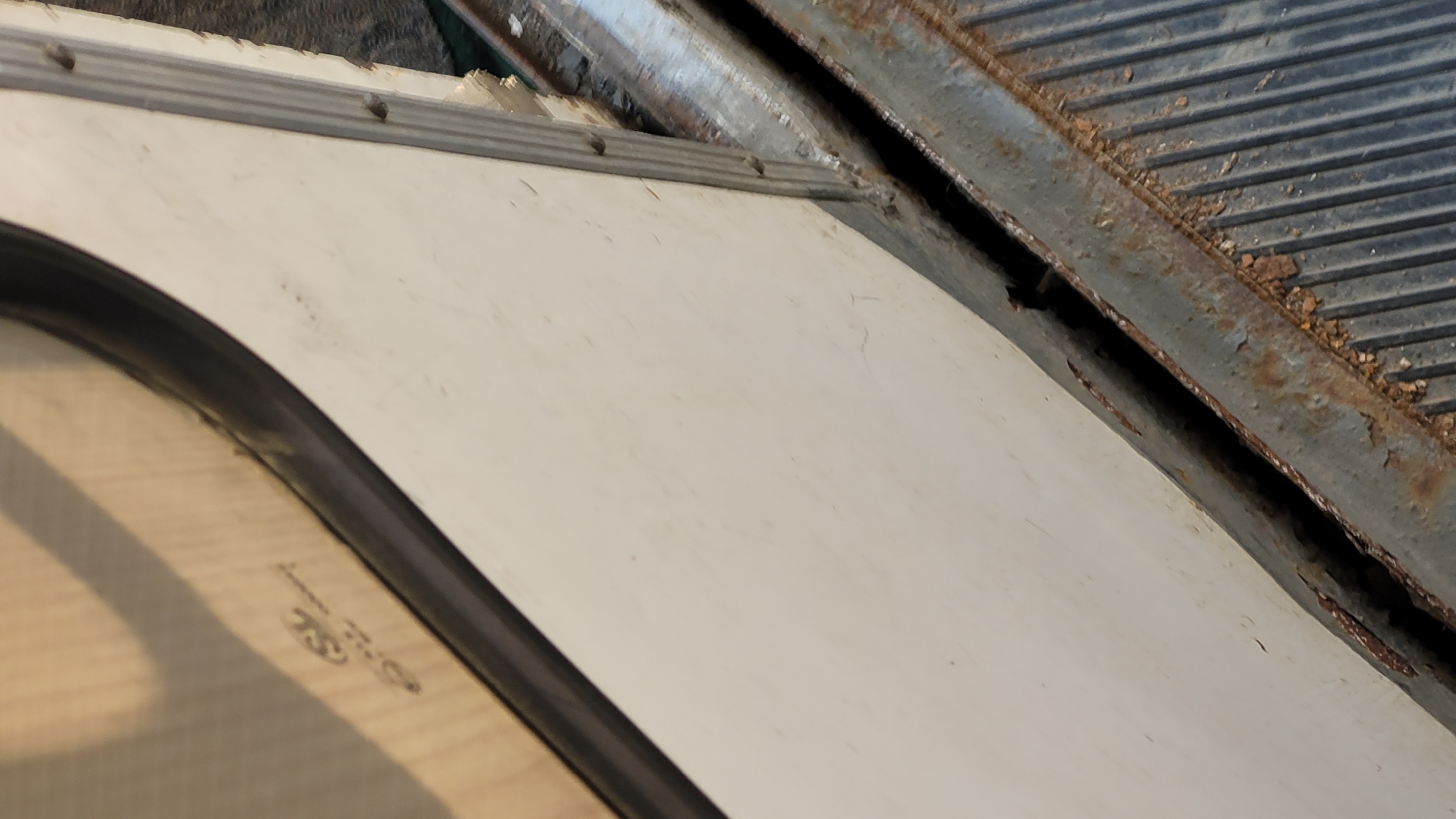 The image shows a section of an unidentified object, possibly a vehicle or structure, situated in an unspecified outdoor environment. The composition is dominated by a curved section of a white, possibly plastic or composite material, panel with a black rubber gasket around a window. Above this panel is a section of rusted metal, featuring a corrugated or grooved surface, indicating age and weathering. The overall impression is one of deterioration and neglect.