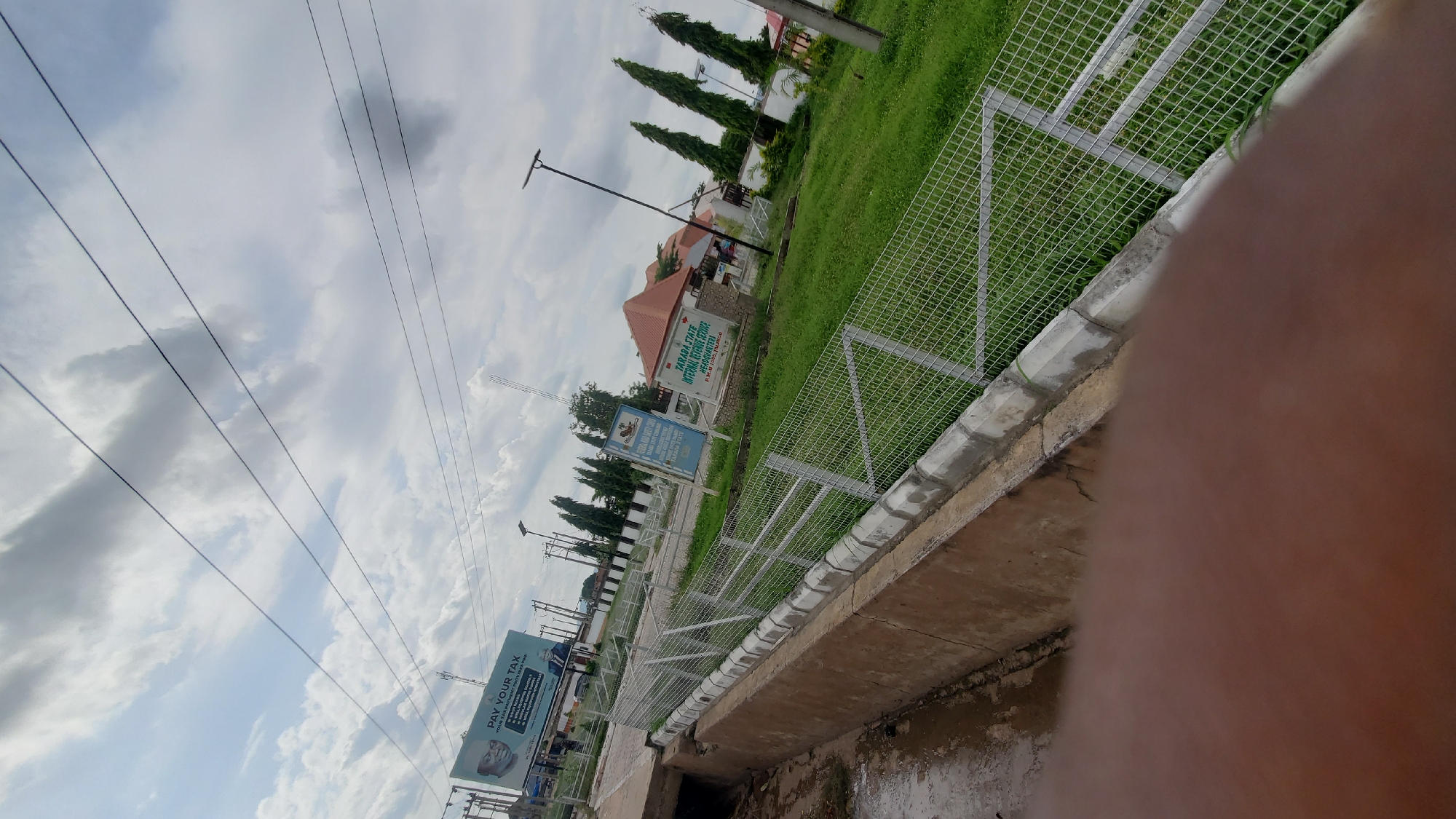 The image depicts a street scene in Jalingo, Nigeria, likely near the Taraba State Internal Revenue Service Headquarters. The foreground is dominated by a concrete drainage ditch filled with murky water and a section of a chain-link fence. Beyond the fence, a well-maintained green lawn stretches towards a cluster of buildings.
On the left side of the frame, a large billboard with the prominent text "PAY YOUR TAX" is visible, featuring a portrait of a man and smaller print detailing various tax-related information. Further down the road, a blue sign with text that appears to be in English and possibly another language is also present.
In the background, several tall, slender trees with dark green foliage are planted in a row. The sky is partly cloudy with a mix of white and gray clouds, and several power lines are strung across the upper portion of the image. The overall impression is one of a functional, administrative area within the city. There are no people clearly visible in the frame.