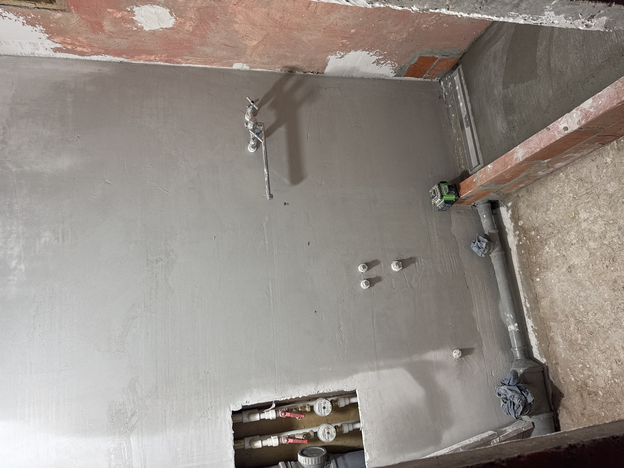 The image depicts an unfinished bathroom in what appears to be an apartment undergoing renovation. The walls are newly plastered with a light gray finish, and the ceiling shows exposed brickwork and remnants of old paint. Plumbing is visible, including water pipes, valves, and a drain pipe wrapped in cloth. A green laser level is positioned on a wooden beam. Located in Chaikovsky, Russia.