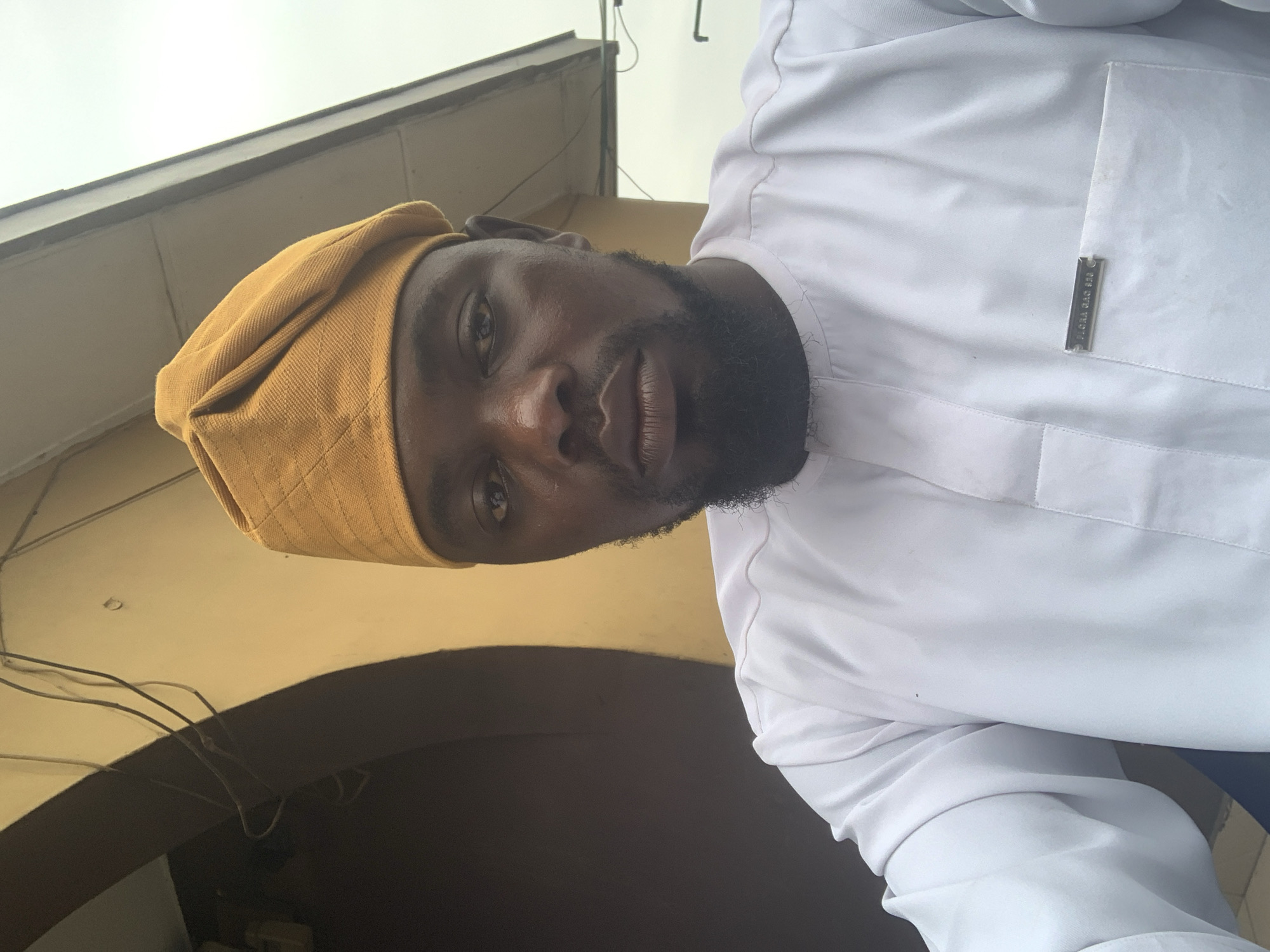 A close-up, eye-level, selfie-style shot shows a Black man wearing a yellow, traditional cap and a white, long-sleeved garment with a Mandarin collar and a chest pocket. The garment has visible stitching details. A metallic badge with an inscription is affixed to the chest pocket. The man has short, dark hair and a beard. He is looking directly at the camera with his mouth closed. The background consists of a textured, pale yellow wall with a dark, arched opening and some electrical wires. Above this, a section of a light-colored overhang is visible against a bright, overcast sky. The scene appears to be outdoors, possibly in an urban setting. Based on visual cues common in Nigerian attire and architecture, the location is likely Shagamu, Nigeria.