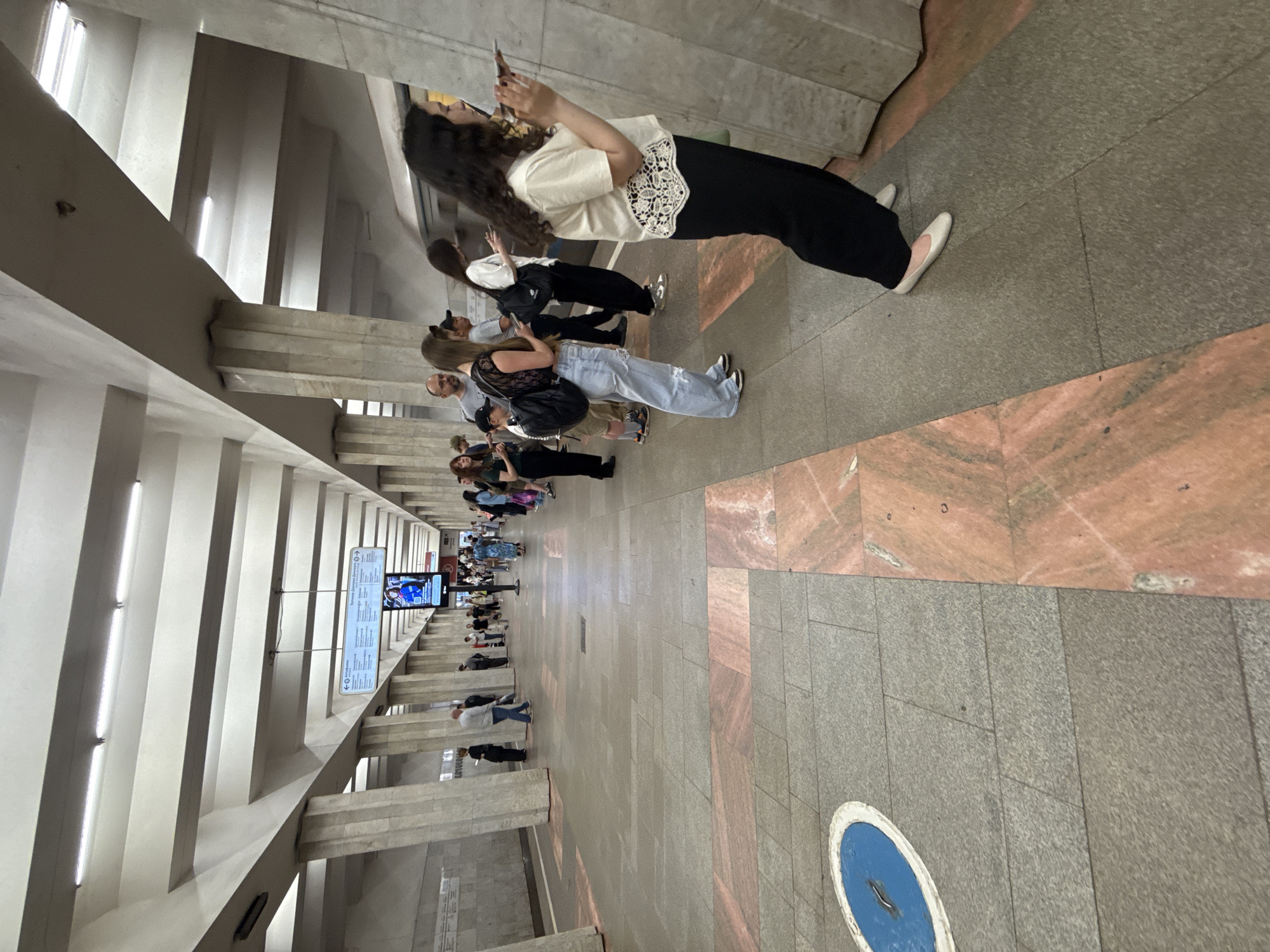 The image depicts a well-lit, spacious metro station in Moscow, Russia. The architecture is characterized by a series of massive concrete columns supporting a ribbed ceiling, creating a sense of grandeur and enclosure. The floor is tiled with large grey and pink granite slabs, with distinct inlaid patterns. A digital display board hangs from the ceiling, providing information about train arrivals and destinations, with a video screen showing a woman.
A diverse group of people are present on the platform, many of whom are standing and looking towards the tracks or interacting with their mobile phones. In the foreground, a woman with long, dark, curly hair is standing on the right side of the frame, facing to the left and looking at her phone. She is wearing a light-colored, short-sleeved top with a white lace-like detail at the hem and black trousers. She has white ballet flats on. Further down the platform, a group of people are lined up, awaiting a train. Among them are individuals with backpacks, casual clothing, and diverse hairstyles. A younger person with light-colored ripped jeans and a black top is visible, as well as a man in a grey t-shirt. The overall atmosphere appears calm and functional, typical of a busy public transportation hub. The presence of the digital display and the general layout suggest it is a modern metro station.