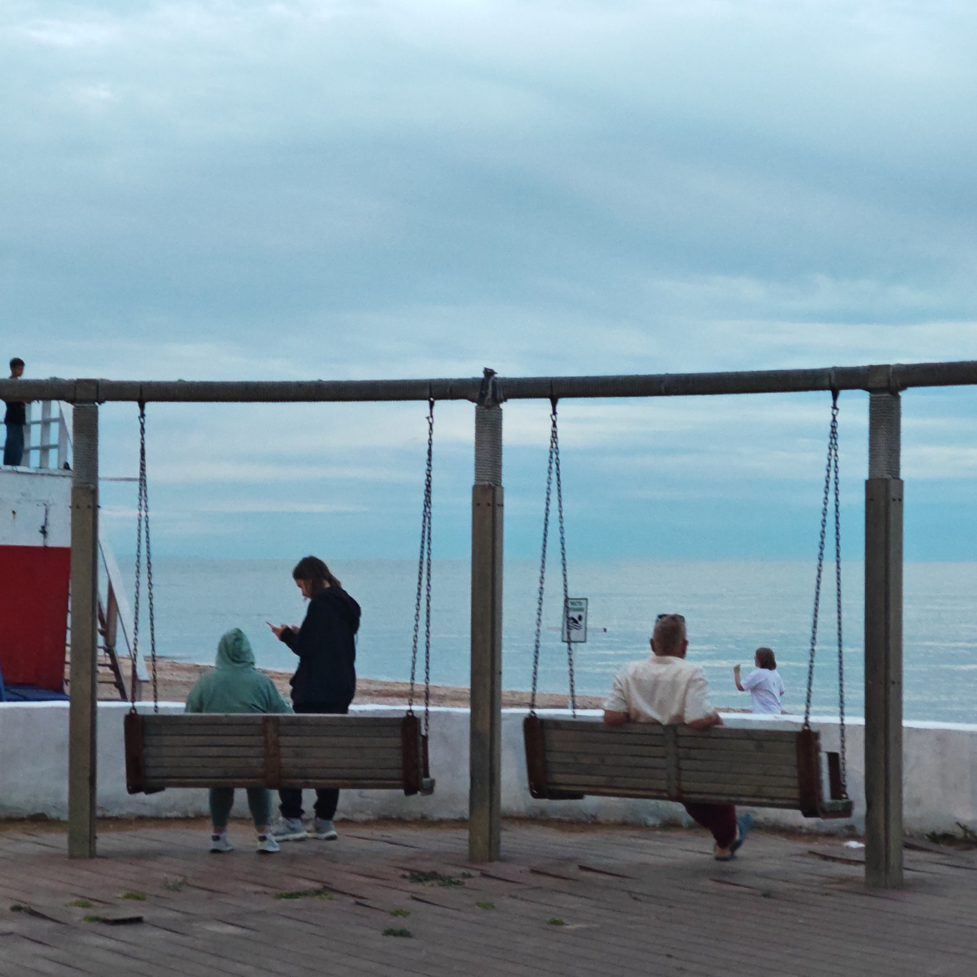 The image captures a coastal scene with several people relaxing by the sea under an overcast sky. In the foreground, a wooden deck supports a large swing structure made of thick metal bars and wooden posts. Two wooden swing benches are suspended by chains from this structure.
On the left swing bench, two individuals are present. One person, wearing a green hooded sweatshirt, sits facing away from the camera, while another, in a dark jacket, is engrossed in their mobile phone. Further left, a person stands on a red and white structure, looking out at the sea.
On the right, a person is seated on another swing bench, facing away from the viewer, dressed in a light-colored shirt and dark trousers or shorts. Beyond the swings, a low white concrete barrier separates the wooden platform from the beach and the calm, blue-grey sea. A distant figure in a white top can be seen near the water's edge in the background.
The sky is uniformly cloudy, casting a soft, diffused light over the scene, suggesting either an overcast day or dusk. A small, blurry sign with an indecipherable icon and text hangs from the swing structure on the right. The setting is a tranquil recreational area by the sea, characteristic of a promenade or pier in городское поселение Щелкино, Ukraine. The general mood is one of quiet relaxation or contemplation.