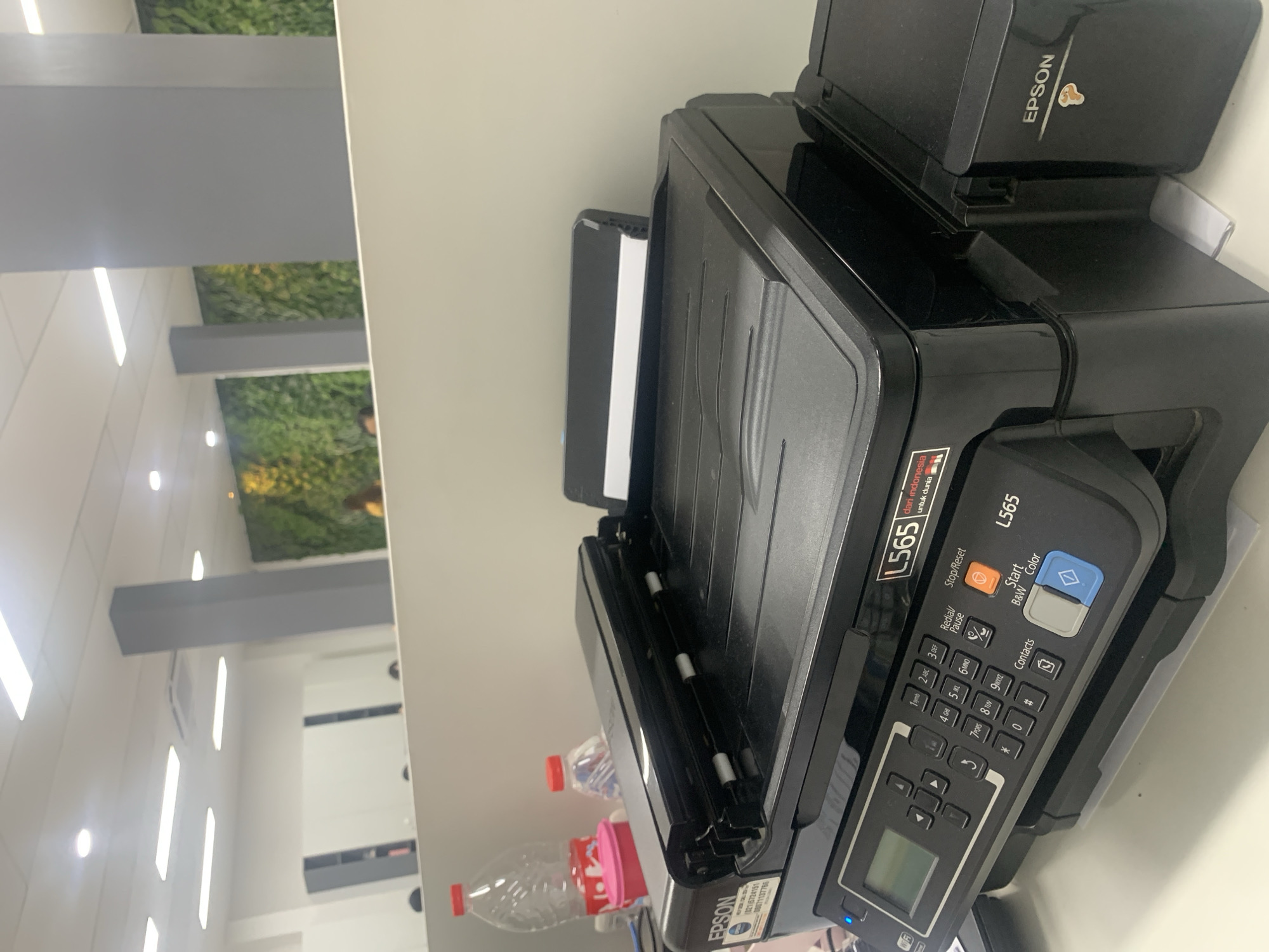 The image shows a black Epson L565 all-in-one printer on a white desk in an office environment. The printer has a scanner on top, a control panel with a small LCD screen and buttons, and an ink tank system on the right side. The background includes office furniture, including grey pillars, white cabinets, and a wall with a vertical green garden feature. A person's head is visible in the far background. There are two plastic water bottles and a pink container on the desk behind the printer. The scene appears to be indoors, likely in an office space in Palembang, Indonesia, as indicated by the text on the printer that reads "L565 dari Indonesia".
