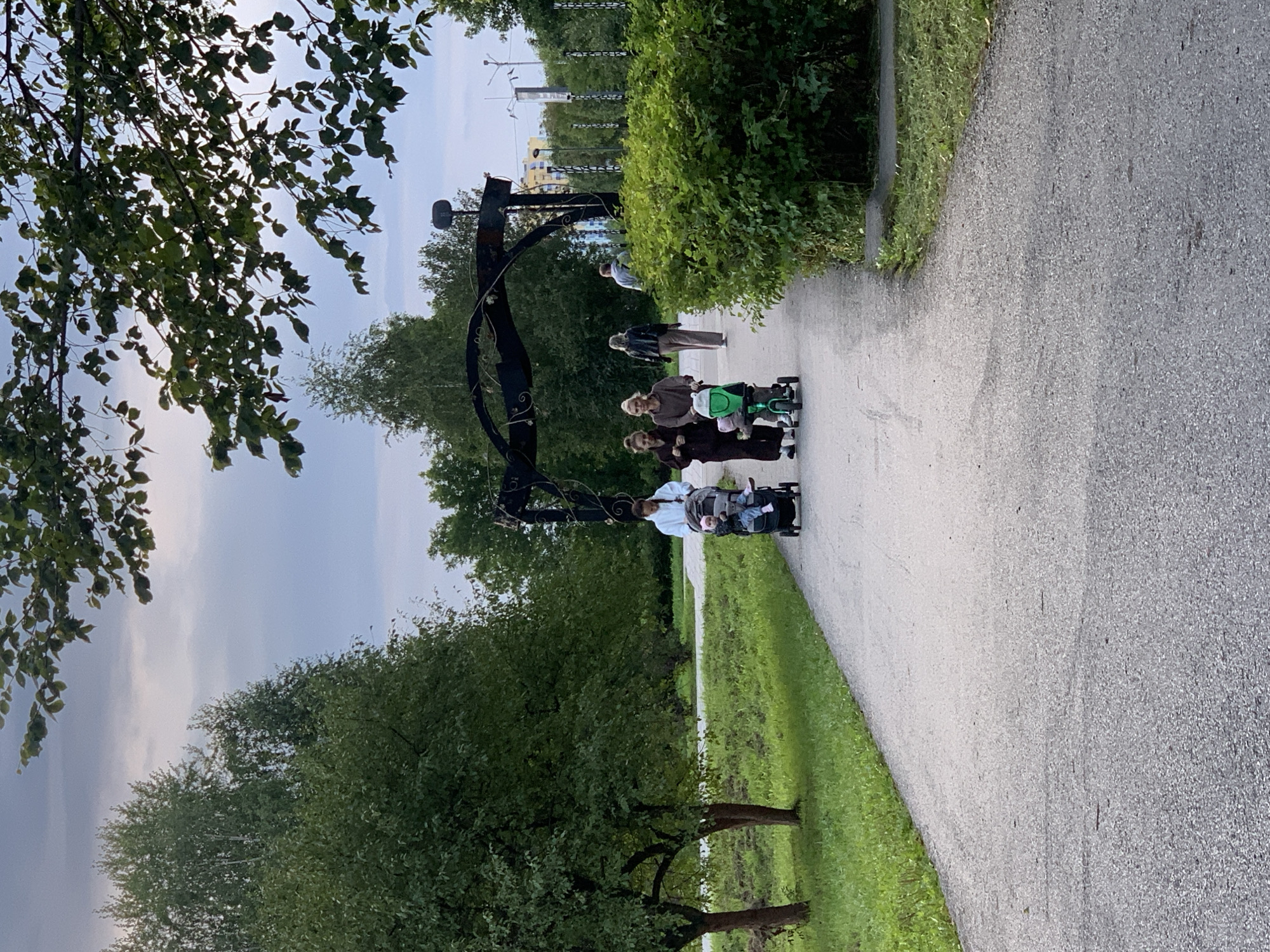 The image depicts an outdoor scene in a park-like setting, featuring a paved pathway winding through grassy areas and trees. Several individuals are visible on the pathway. In the foreground, two women are walking with strollers. One woman is pushing a stroller with a child in it, while the other woman walks alongside her. Further back, another woman walks behind them, and a man is seen walking in the distance. An elaborate metal archway is visible in the background, spanning over the pathway. The environment appears to be a public park with lush green trees and bushes. The sky is overcast, suggesting it might be a cloudy day. There is no specific information within the image to indicate its location in Prokopyevsk, Russia, or any other specific city.