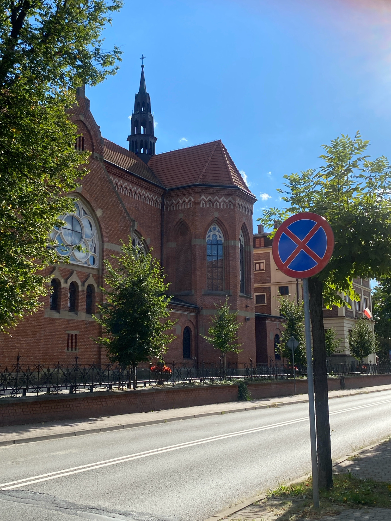 The image shows a brick church with a tall steeple and large stained-glass windows. A no-parking sign is visible in the foreground, near a tree. A road runs in front of the church, with a sidewalk and a low brick wall topped with an ornate black metal fence. The sky is clear and blue, with sunlight glinting from the top right. The architectural style of the church suggests it is likely located in Central Europe, possibly Poland. Further investigation of the church's architecture, the style of the no-parking sign, and any visible text or flags would be necessary to confirm the precise location within Nowy Sącz, Poland.