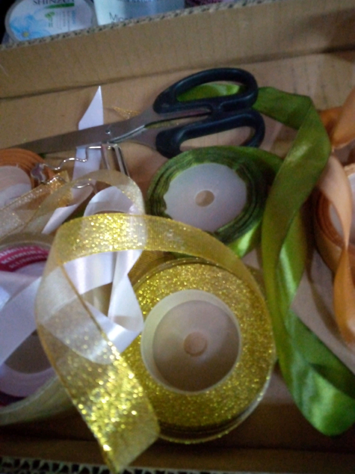 A close-up, overhead shot captures a variety of colorful ribbons, some rolled and some loose, along with a pair of black-handled scissors, all contained within a cardboard box. The ribbons are in shades of gold, green, white, and orange, with some featuring a glittery texture. The scissors are positioned on top of the ribbons, appearing to be in use or ready for use. The box itself is made of corrugated cardboard. The scene is consistent with a crafting or gift-wrapping area. The specific location in Sumberwono, Indonesia is not discernible from the image.