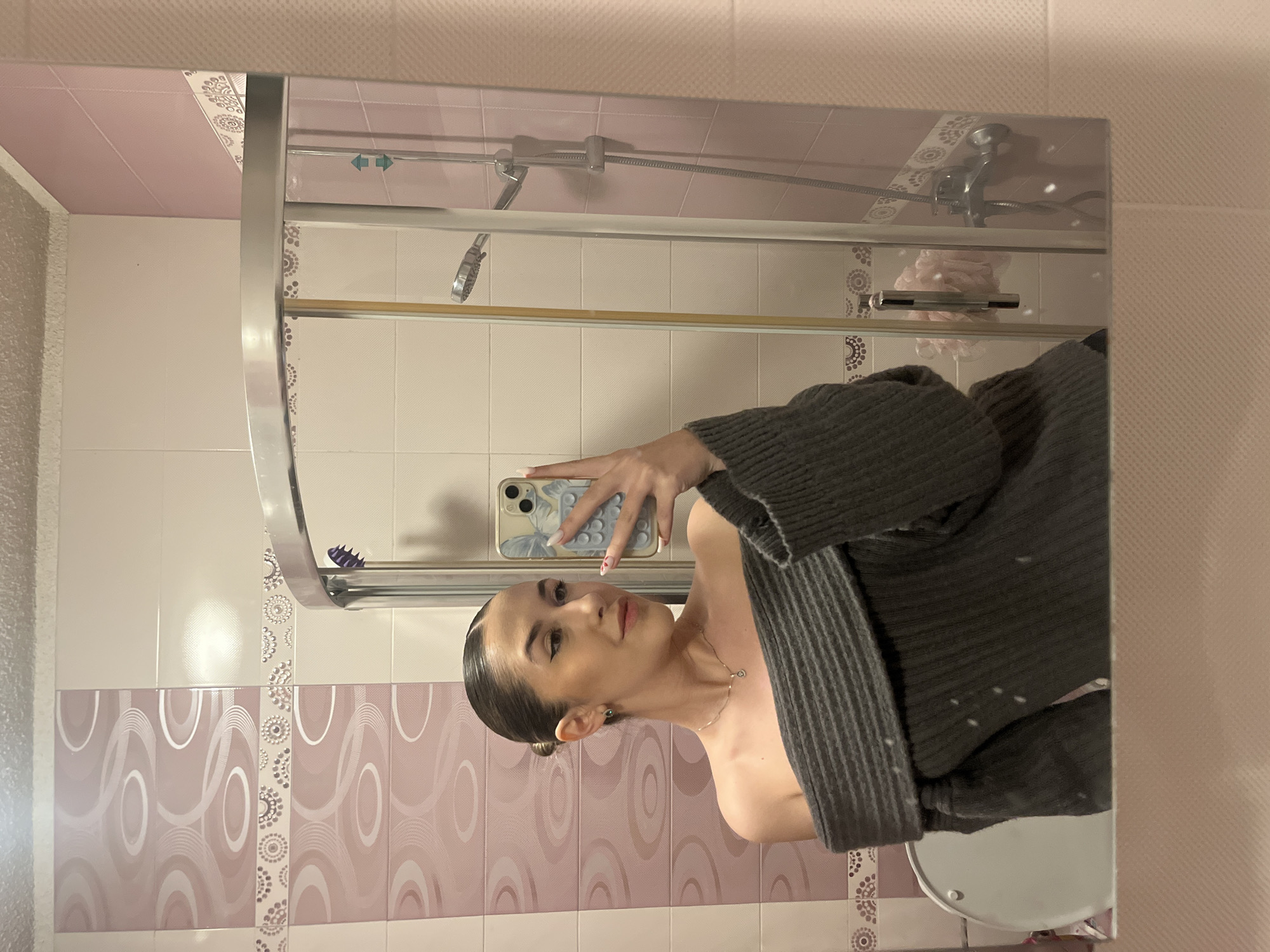 A woman is taking a selfie in a bathroom. The bathroom has a shower with a glass door, pink and white tiled walls, and a pink ceiling. The woman is wearing a gray sweater and holding a smartphone with a butterfly-patterned case. The reflection shows a sink. The location cannot be determined from the provided information.