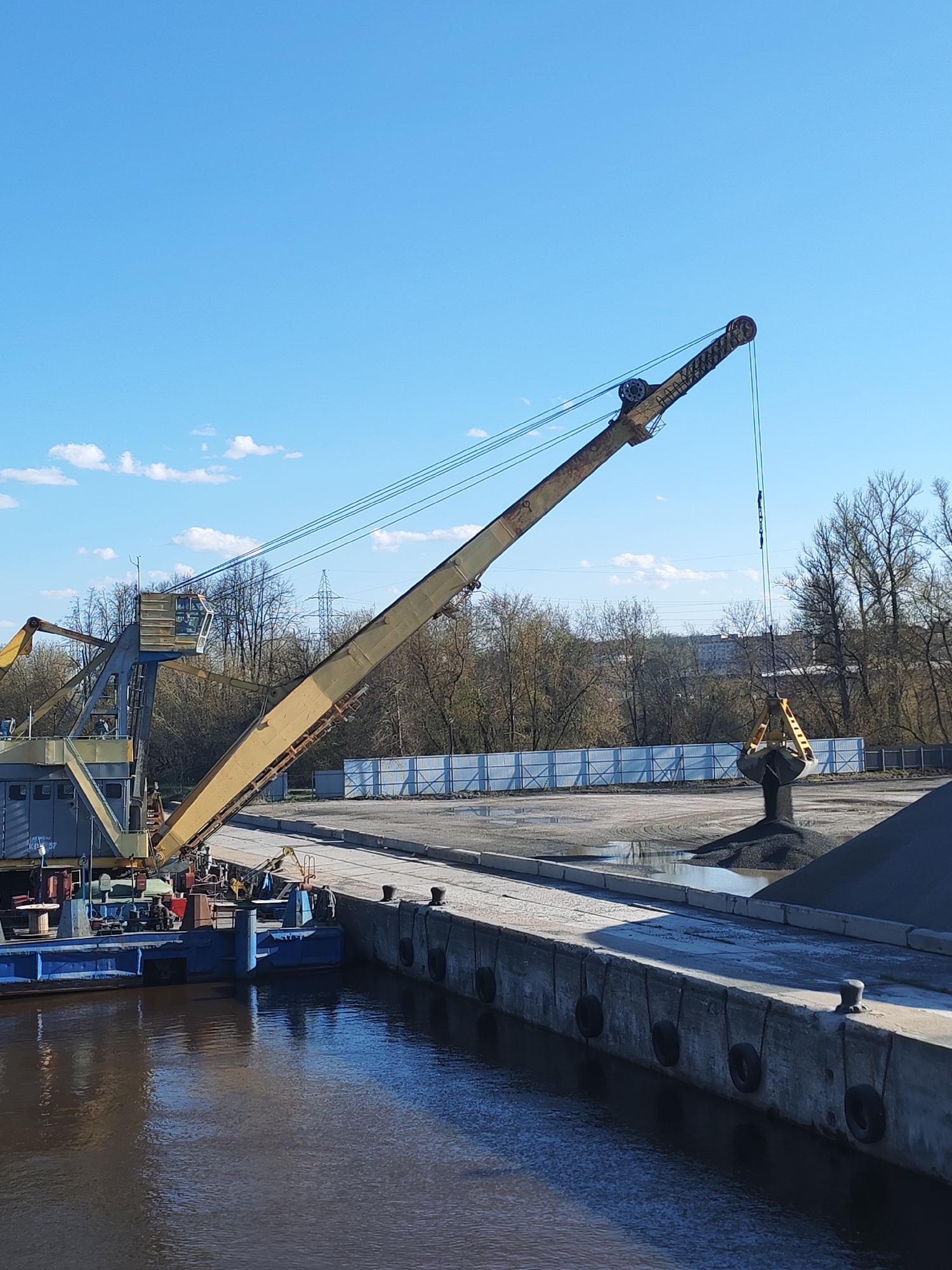 The image depicts a dredging crane positioned on a barge, located at a dock along a waterway. The crane is actively unloading a pile of dark material, possibly gravel or coal, onto the dock. A white fence runs along the background of the dock area. The water is calm, reflecting the sky and the barge. The sky is blue with scattered clouds. Bare trees can be seen behind the fence. The location is identified as Yakhroma, Russia.