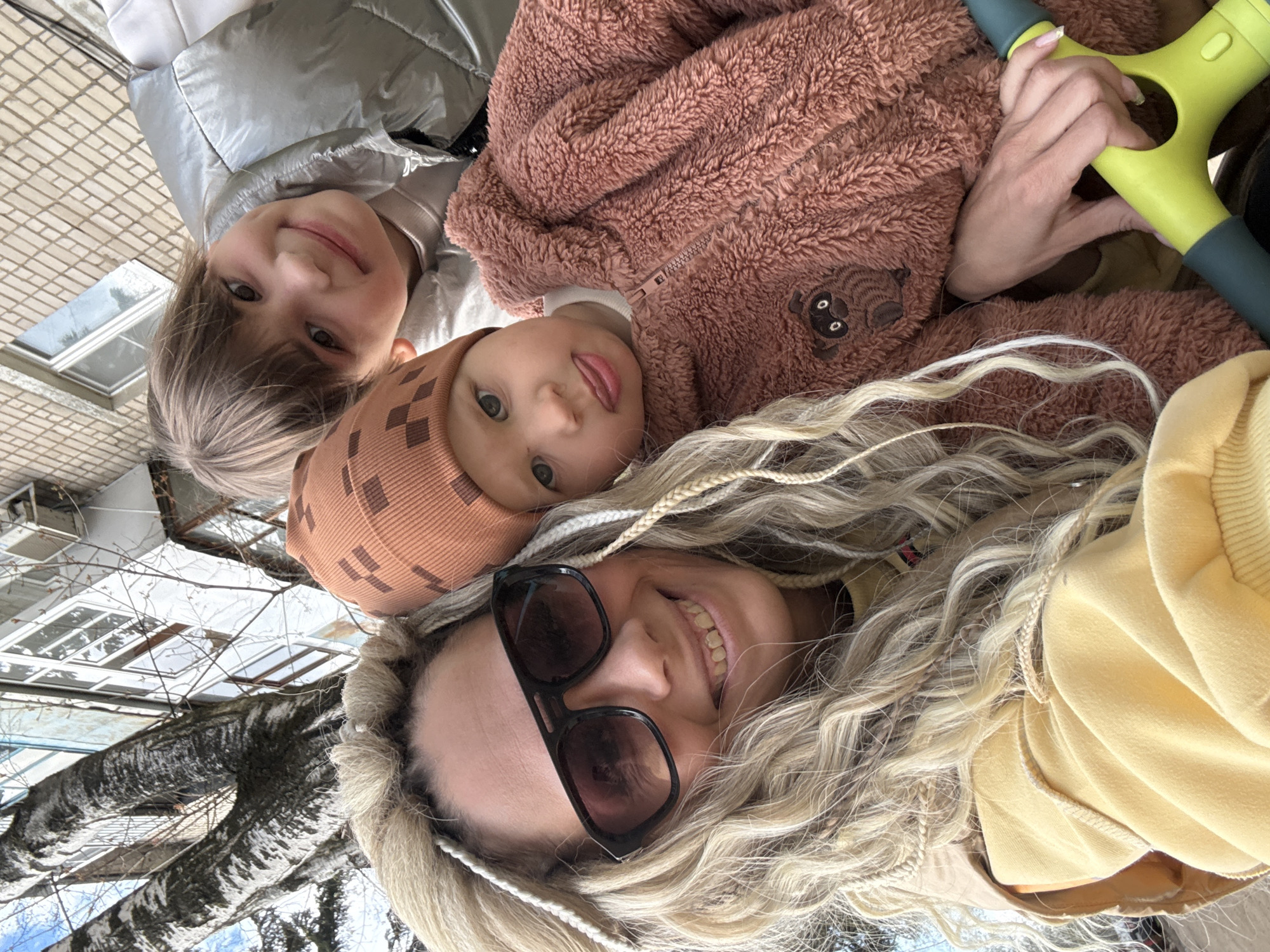 The image shows a woman and two children lying on what appears to be a paved surface, likely a sidewalk or plaza. The woman, positioned at the bottom of the frame, has blonde hair styled in braids, is wearing sunglasses, and is smiling. Above her is a young child wearing a brown patterned hat and a pink teddy bear jumpsuit. Above that child is a slightly older child wearing a silver puffer jacket, also smiling. The background features buildings with windows and bare trees. The scene is likely set outdoors during daylight in the city of Rusanovo, Russia. One of the children is holding a green plastic toy.